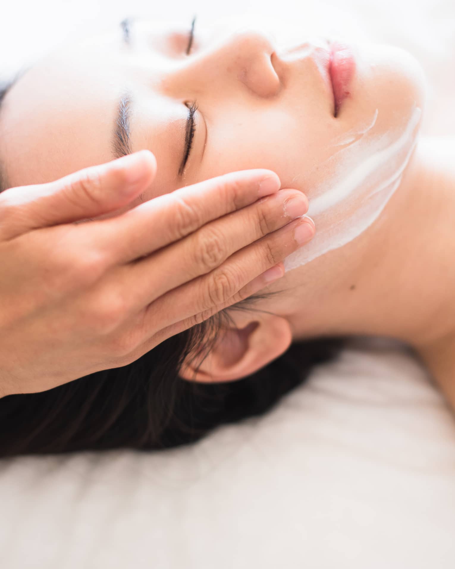 Close-up of spa attendant's hands rubbing white cream on woman's face as she lies on massage table