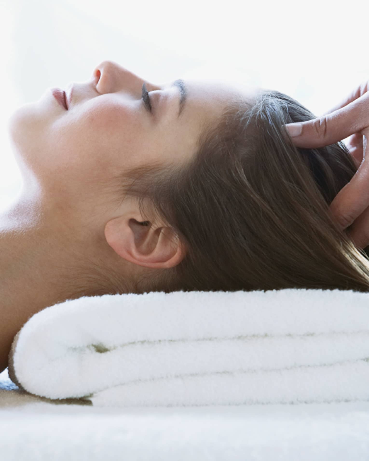 Woman lays on back with head on folded white towel, closes eyes as hands massage her scalp