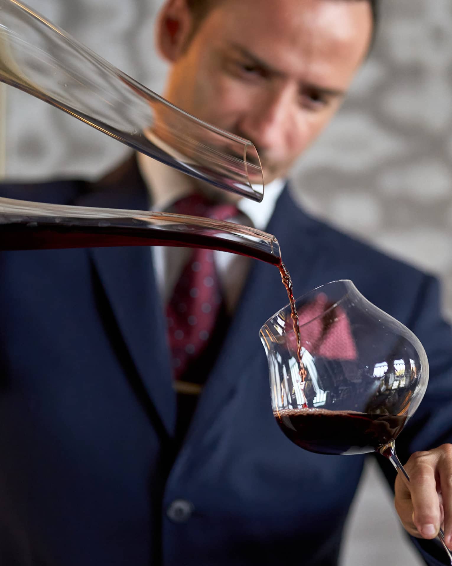 Male in dark blue suit pours a glass of red wine from a large decanter