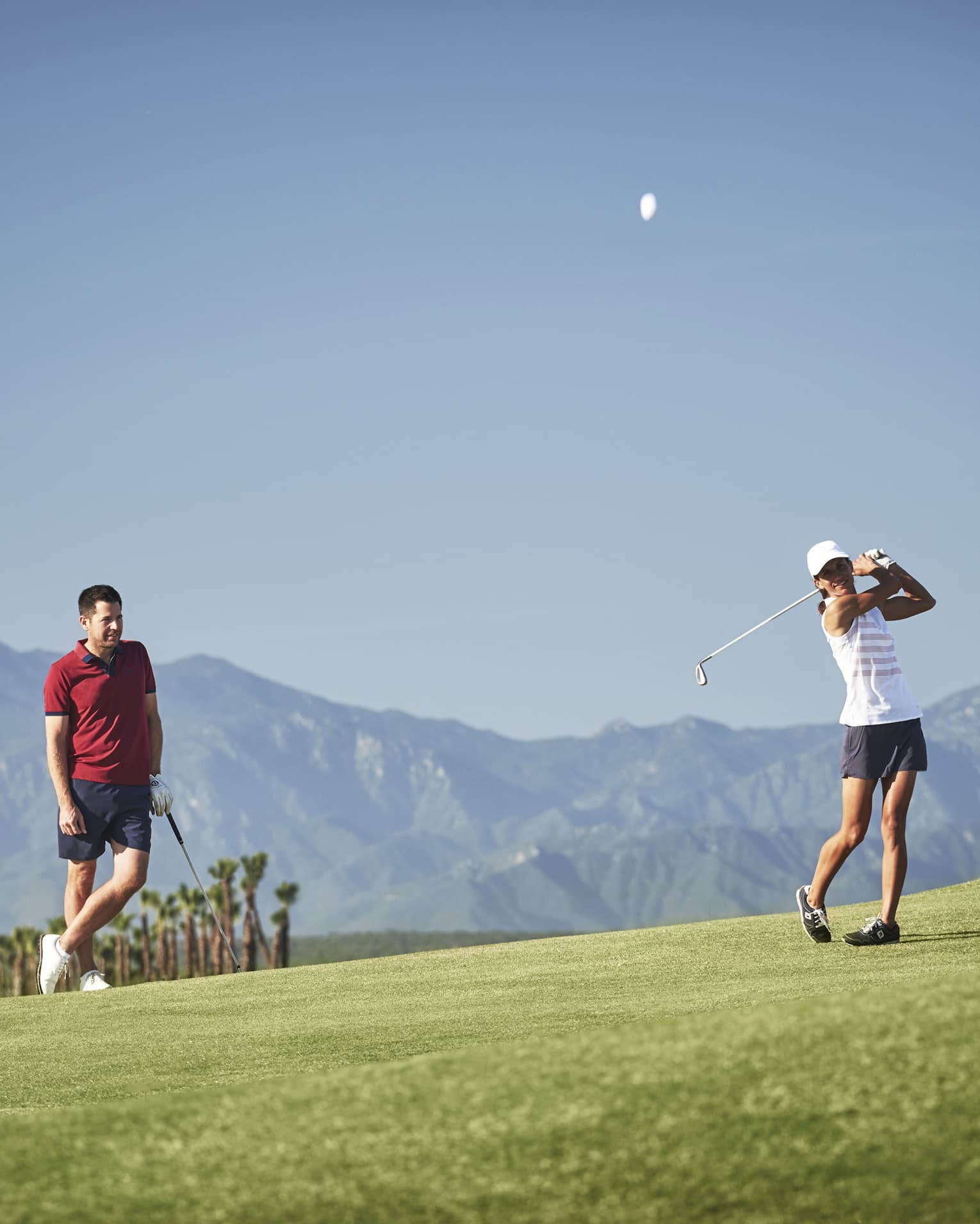 A golfer leans on their club observing another's swing on a picturesque golf course silhouetted by a mountain range.