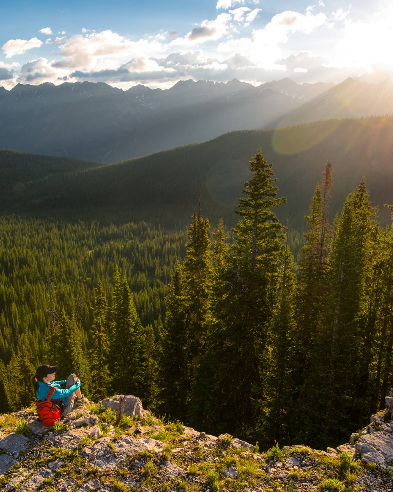 Person sits at edge of cliff overlooking trees, mountains as sun starts to set