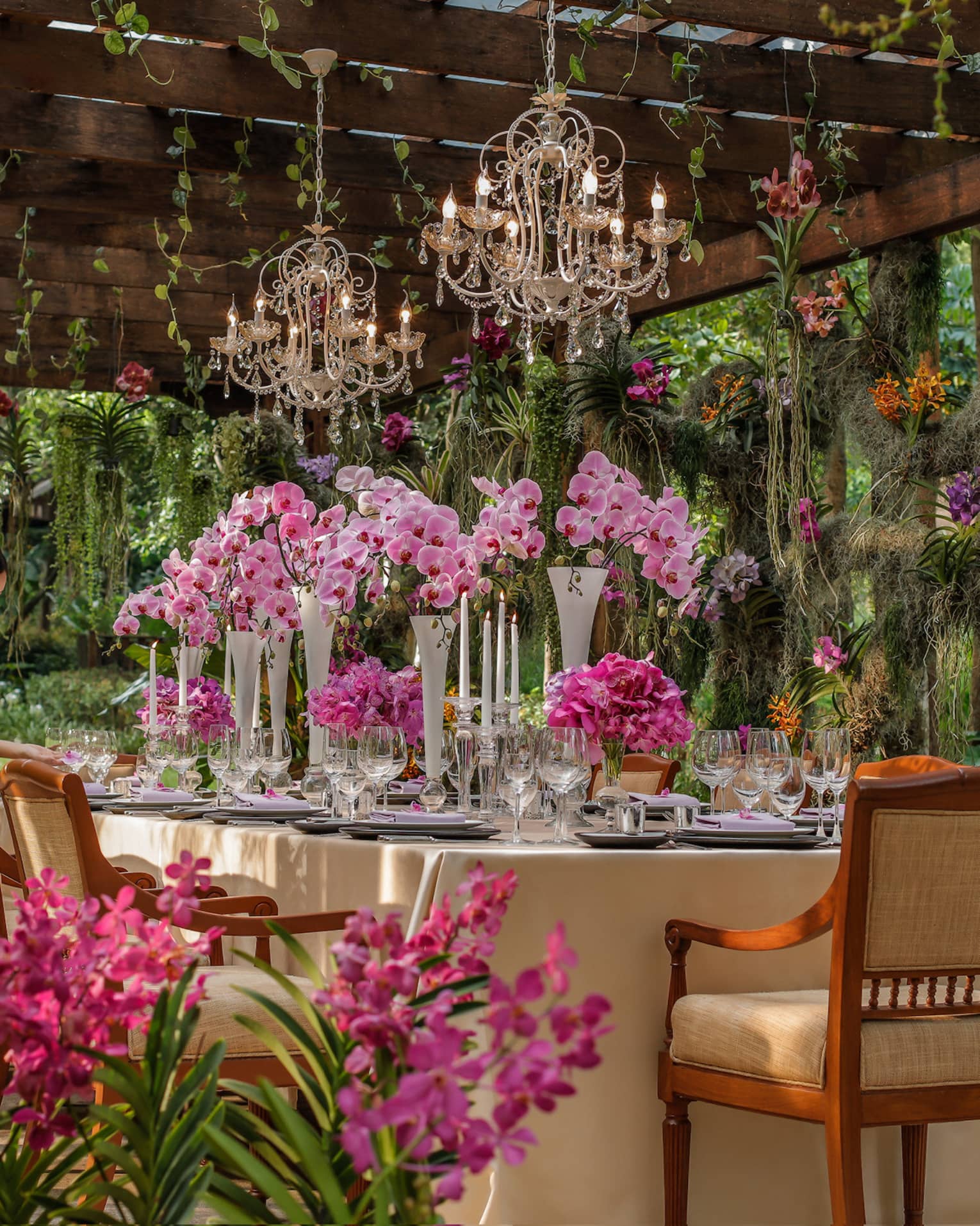 Hotel staff sets large formal dining table in tropical garden, large pink flowers under small crystal chandeliers