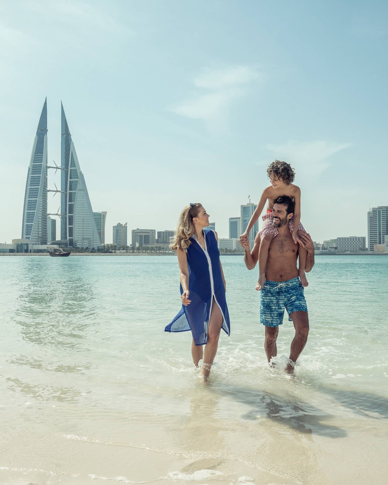 A man with a teenager on his shoulders walks with a woman through shallow ocean water with the city of Bahrain Bay in the background 