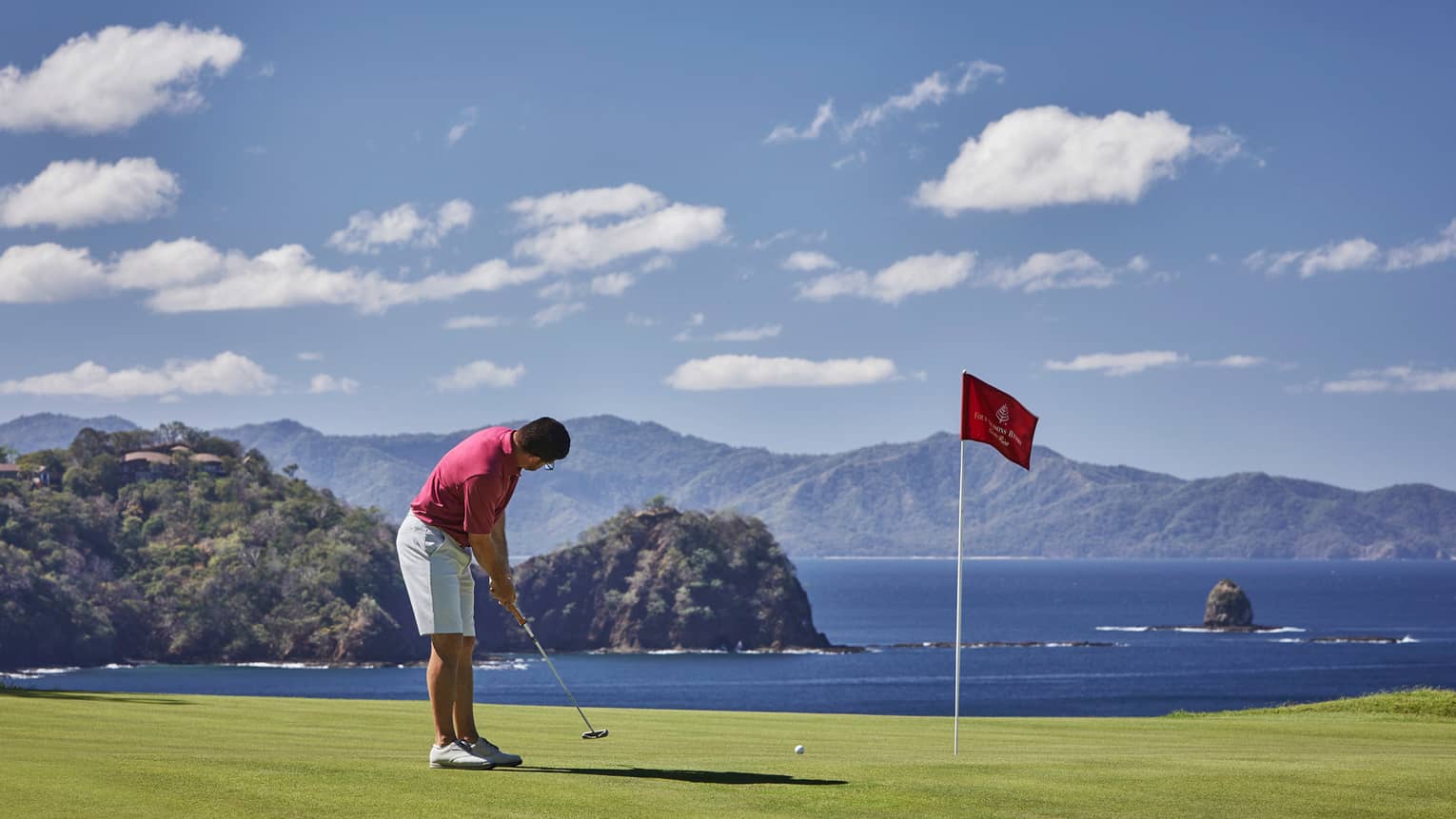 Man prepares to putt near red flag on golf green overlooking ocean 