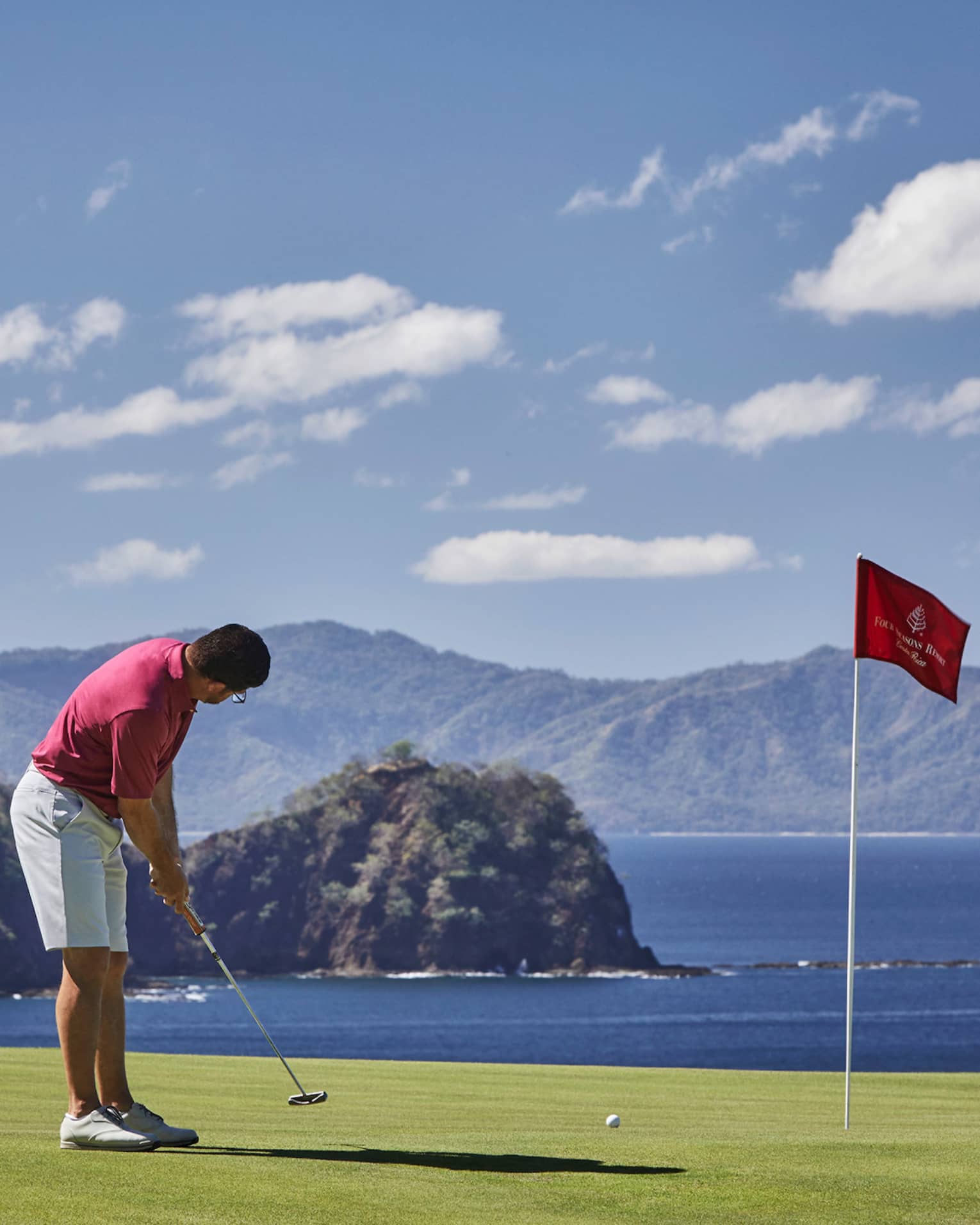 Man prepares to putt near red flag on golf green overlooking ocean 