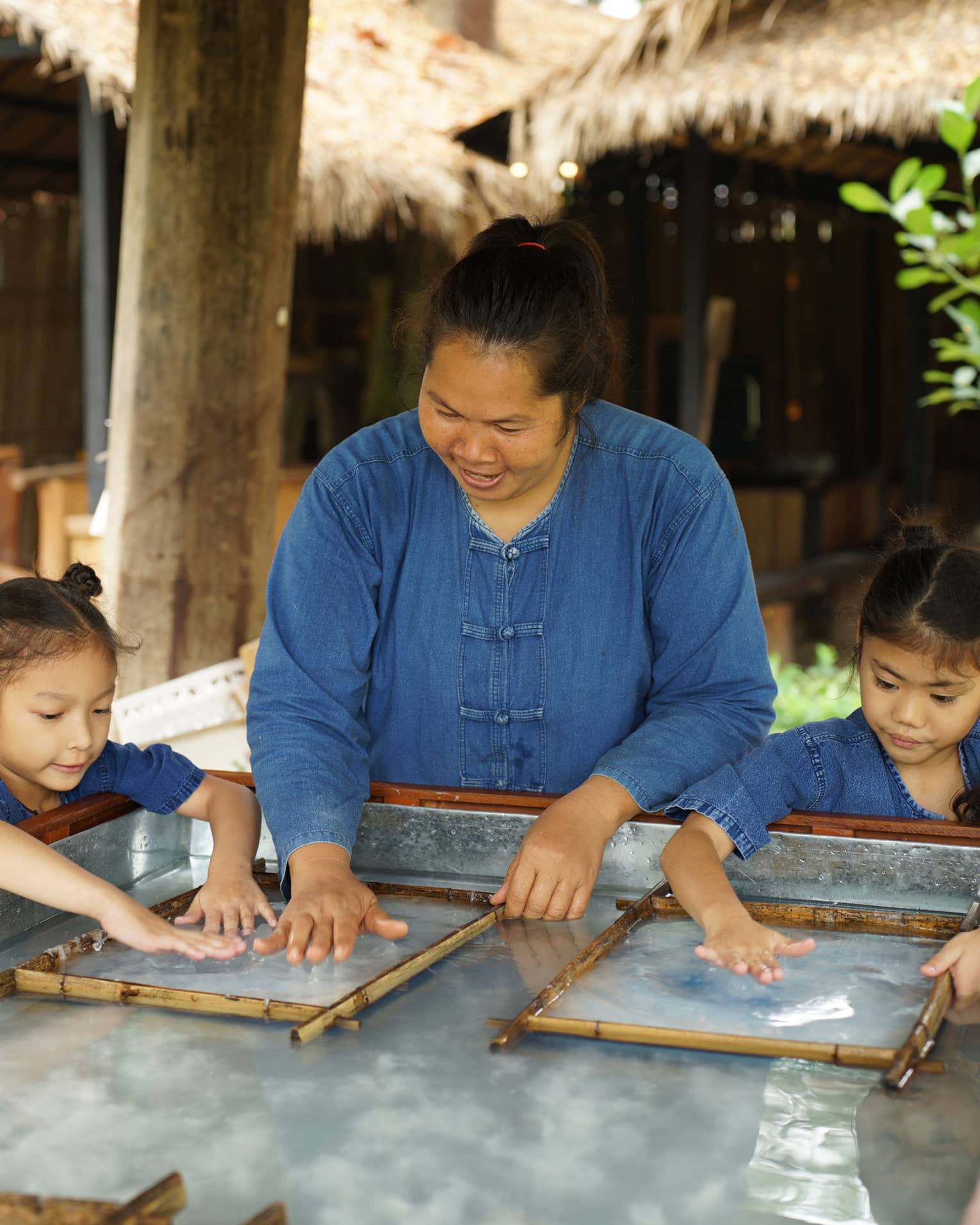 An adult stands between two children, running their hands along a clear, wet substance enclosed in two thin bamboo frames.