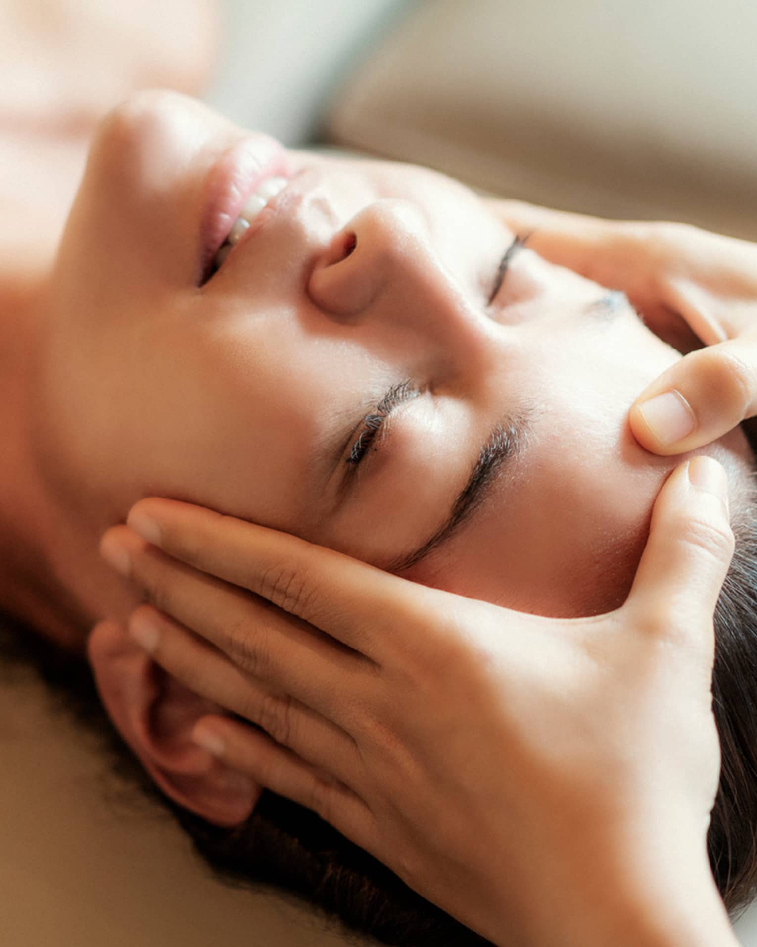 Spa facial, two hands rest on woman's forehead as she closes her eyes, lays under sheet on treatment table