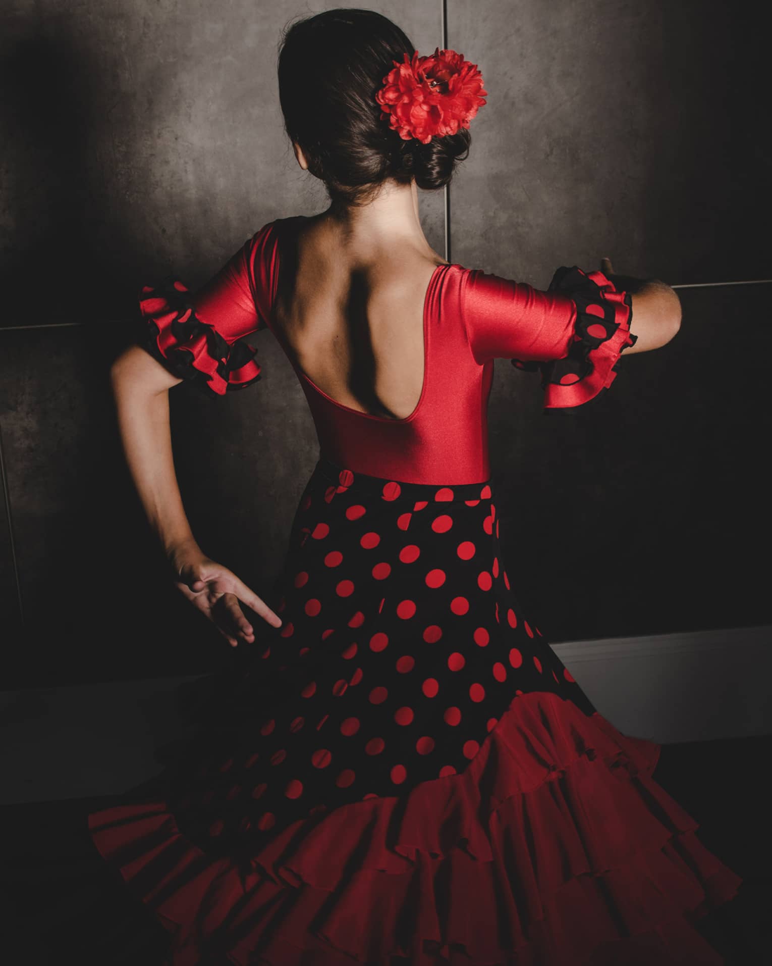 A woman wearing a black and red flamenco dress and red flower in her hair dances with her face turned away from the camera 