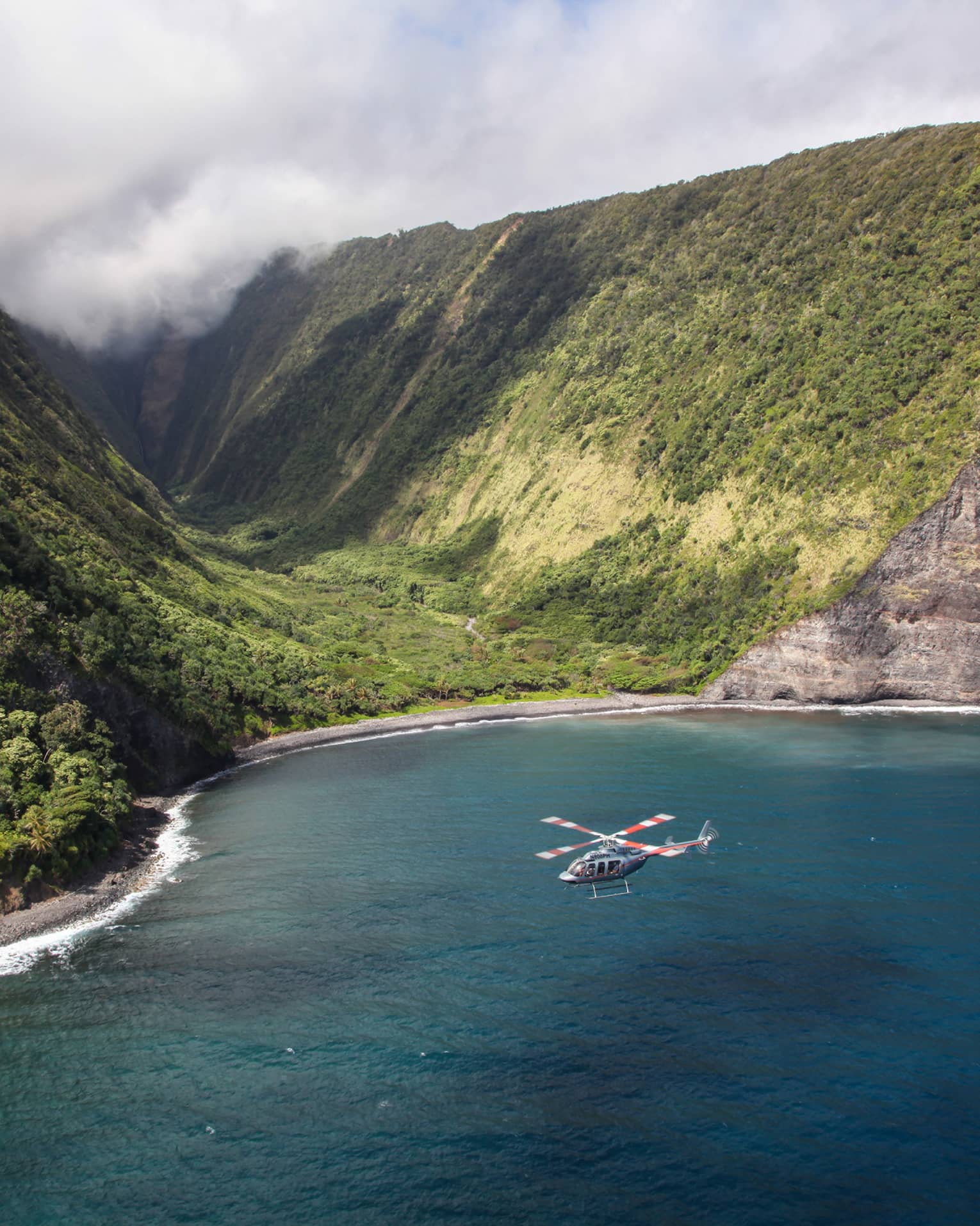 Helicopter flies over ocean by sweeping green volcanic mountains