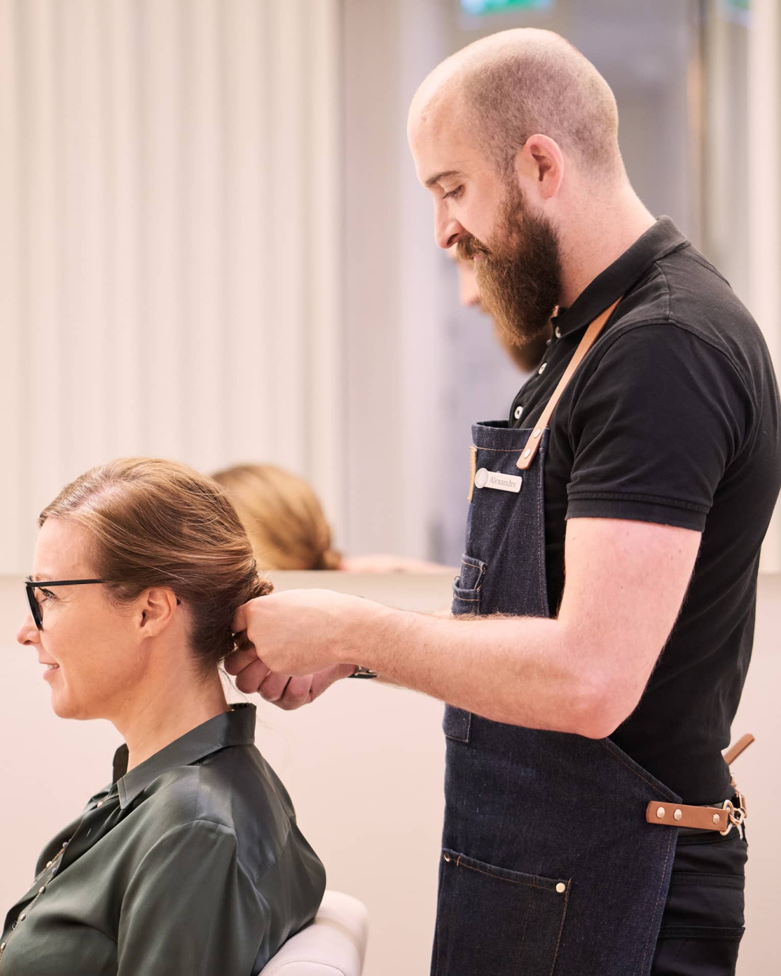 Woman sits in salon chair as hair stylist with long beard holds her hair