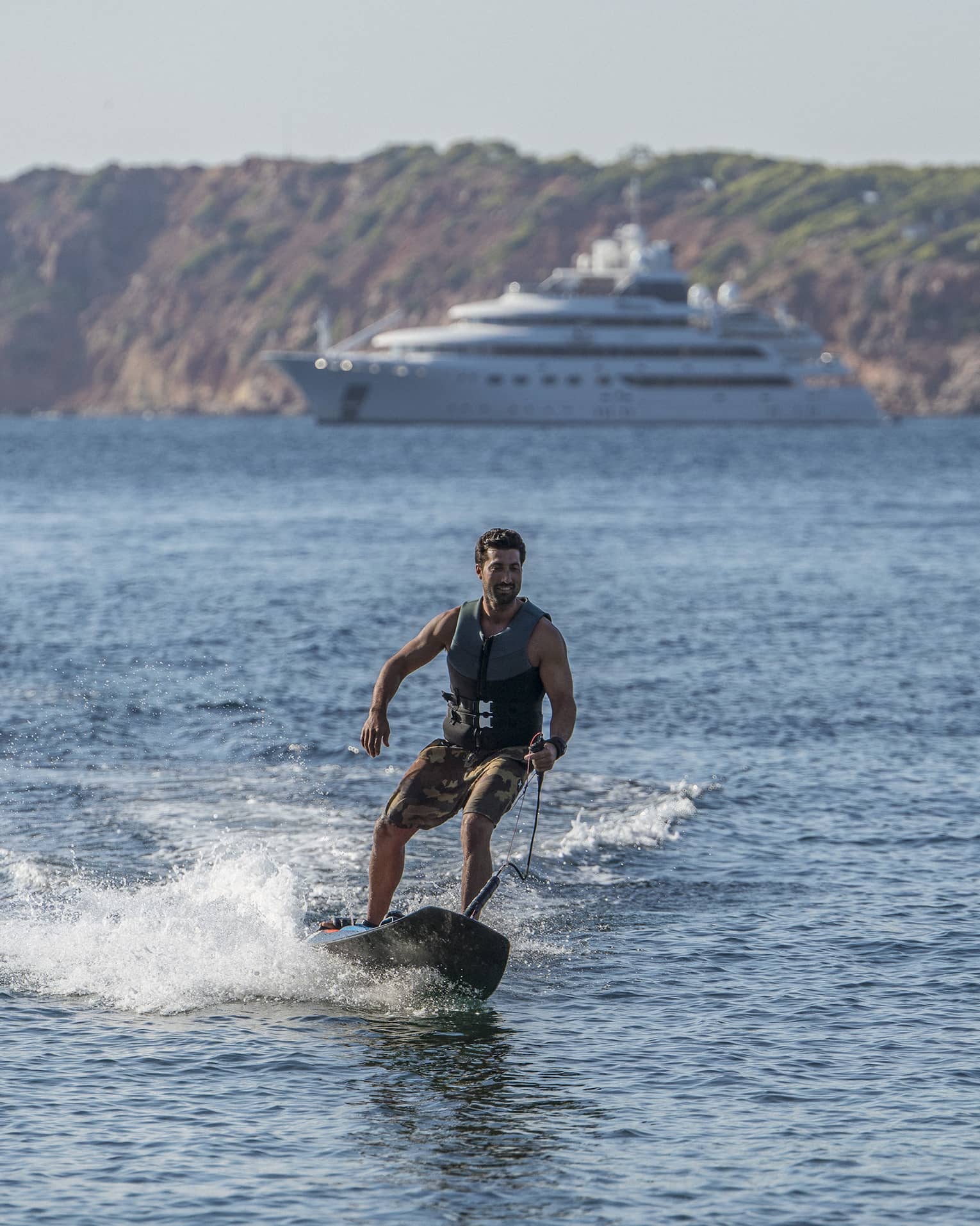 Front view of a guest jetting along the sea on a motorized surfboard, a majestic ship and forested mountains in the backdrop.