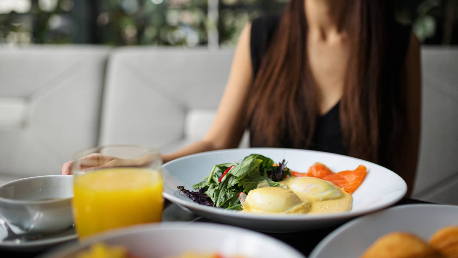 A plate of eggs benedict and a glass of orange juice sit on a table in front of a woman