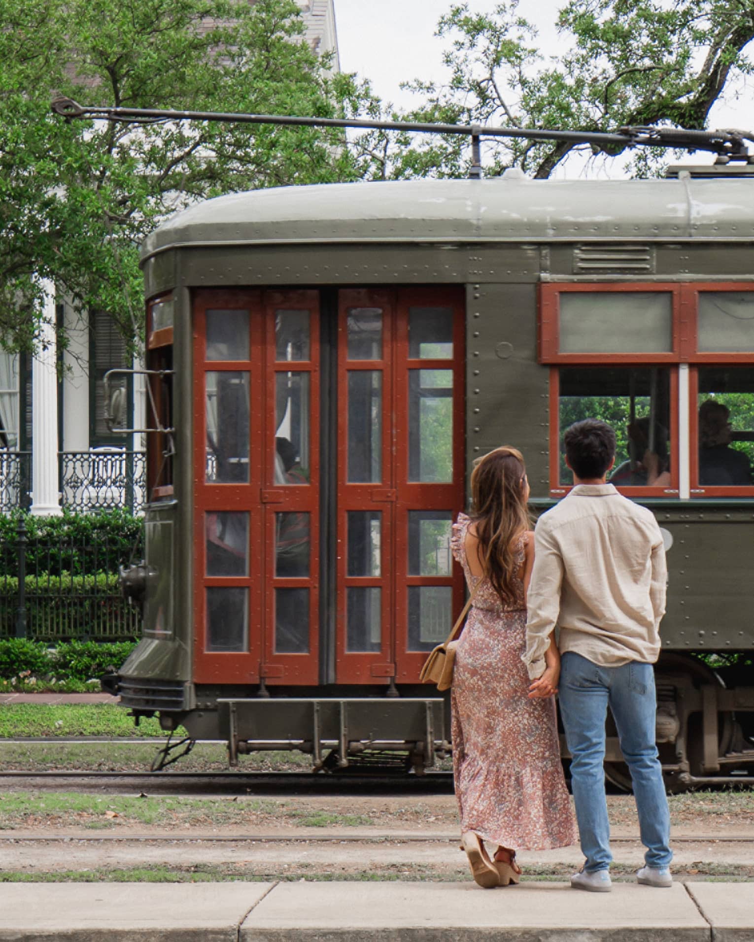 Woman and man hold hands in front of old-fashioned streetcar on New Orleans street, white house in background