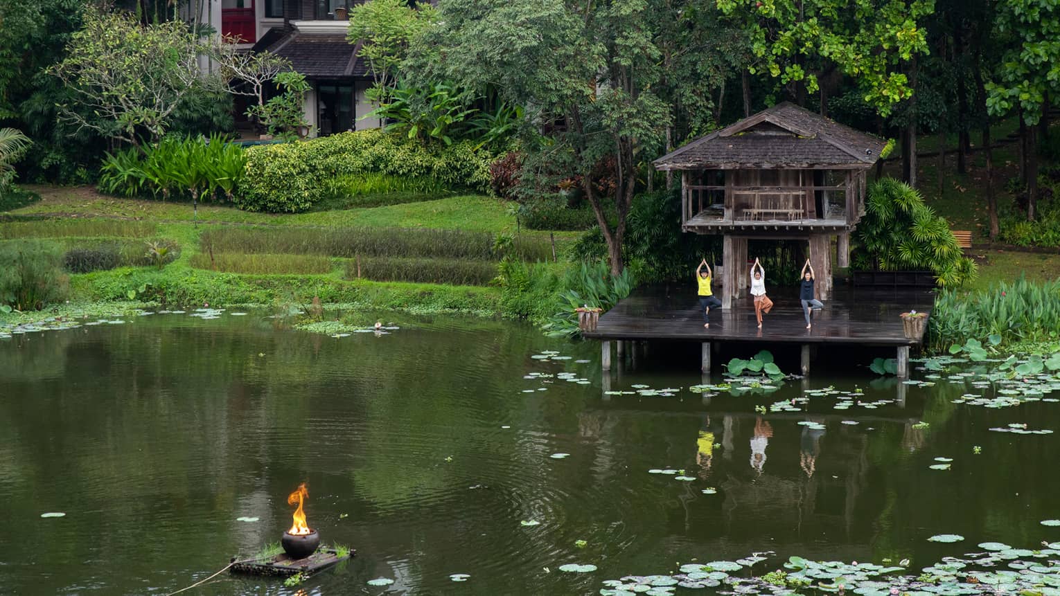 A family does yoga on the porch of a brown building, overlooking a green pond covered in lilypads.