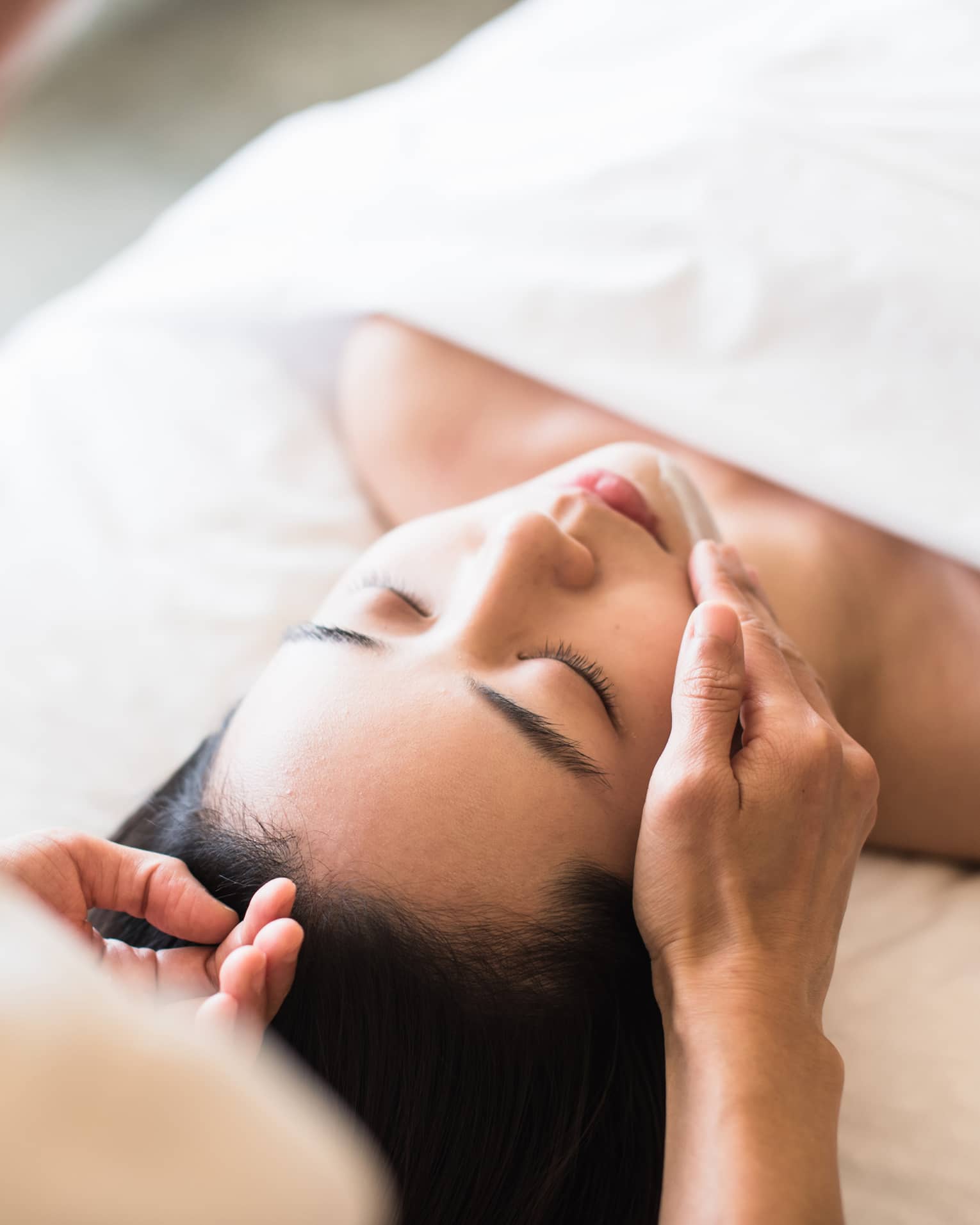 Woman lying under white sheet on massage table, eyes closed as spa staff rubs lotion on her chin