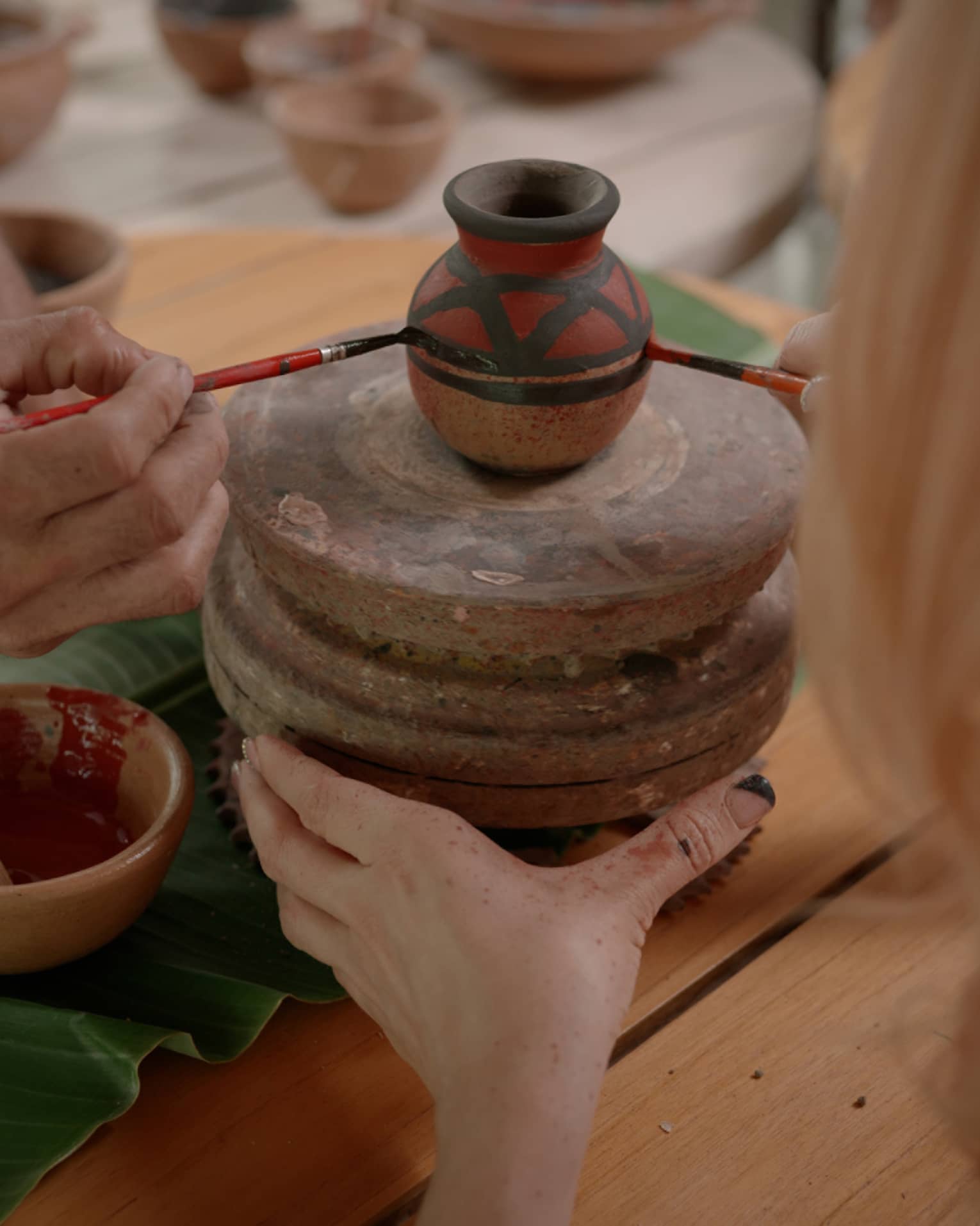 Two sets of hands using thin paintbrushes to decorate a small ceramic vase on a pottery wheel amid pots of paint and pigment.