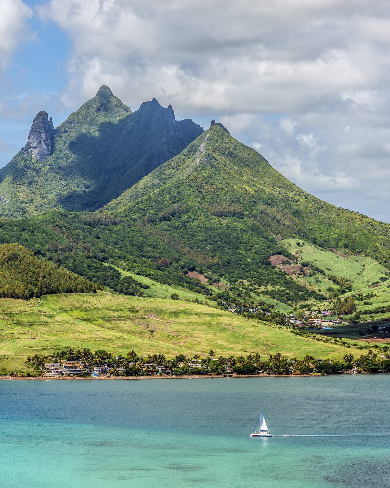 Island of lush, forested, multi-hued jagged hills over turquoise waters, a sailboat in the foreground dwarfed by the hills.