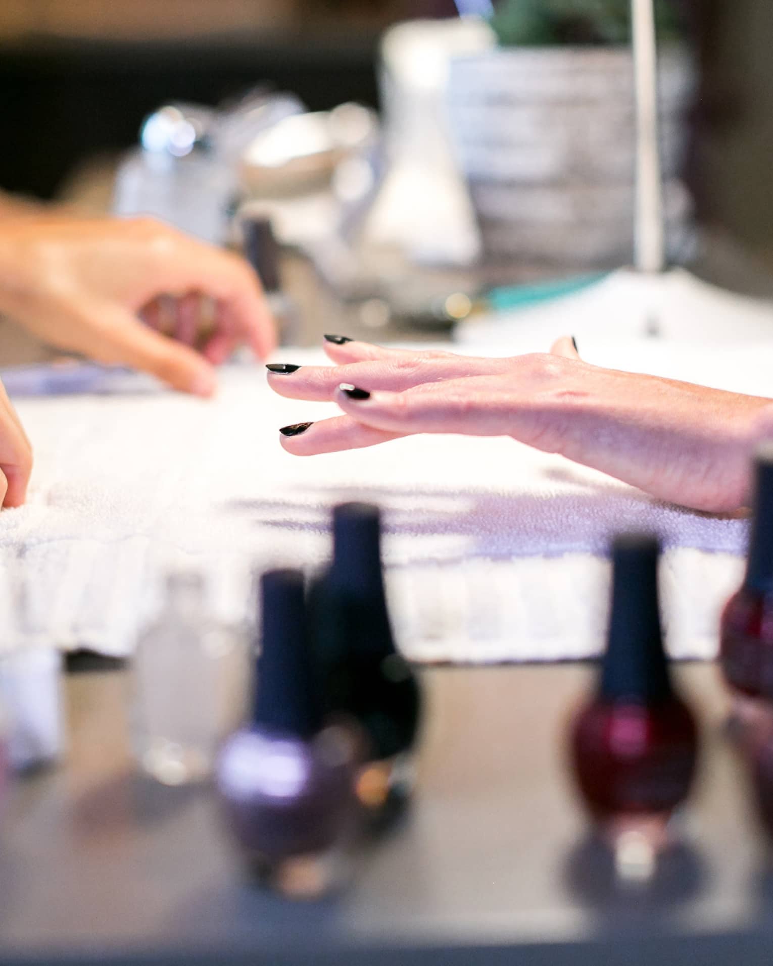 Close-up of woman's freshly-painted nails in dark colour above white cloth, nail polish bottles in foreground