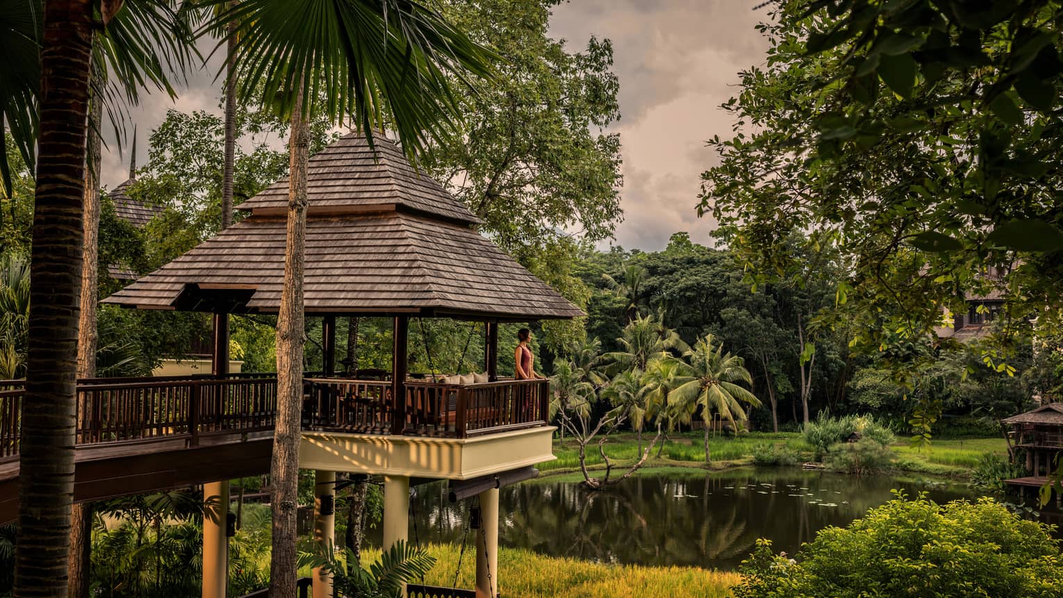 Woman stands at edge of balcony under shingled-roof, overlooking pond and tropical gardens