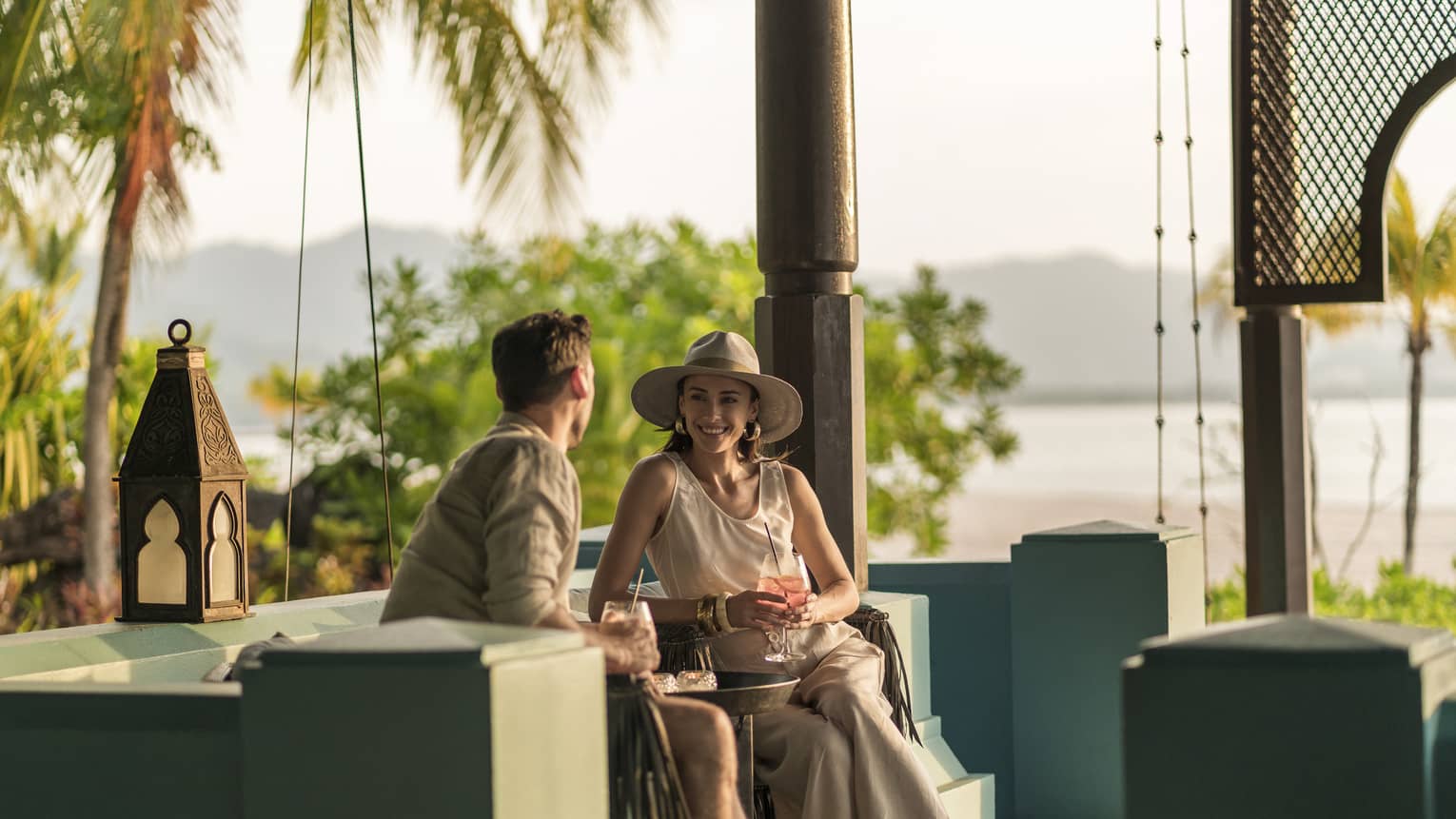 A man and woman are sitting in an outdoor Rhu Bar area overlooking the ocean holding cocktails