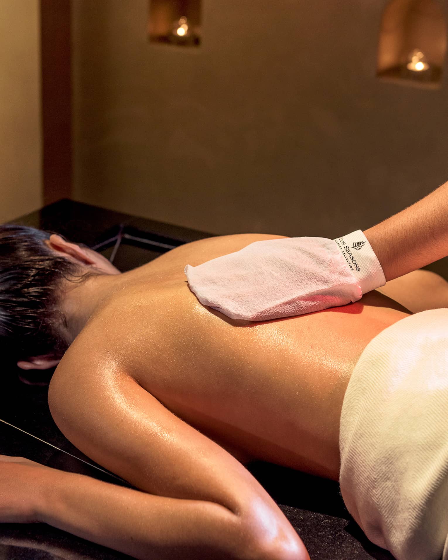 Woman lies face-down on spa table while hand with white mitten massages her back