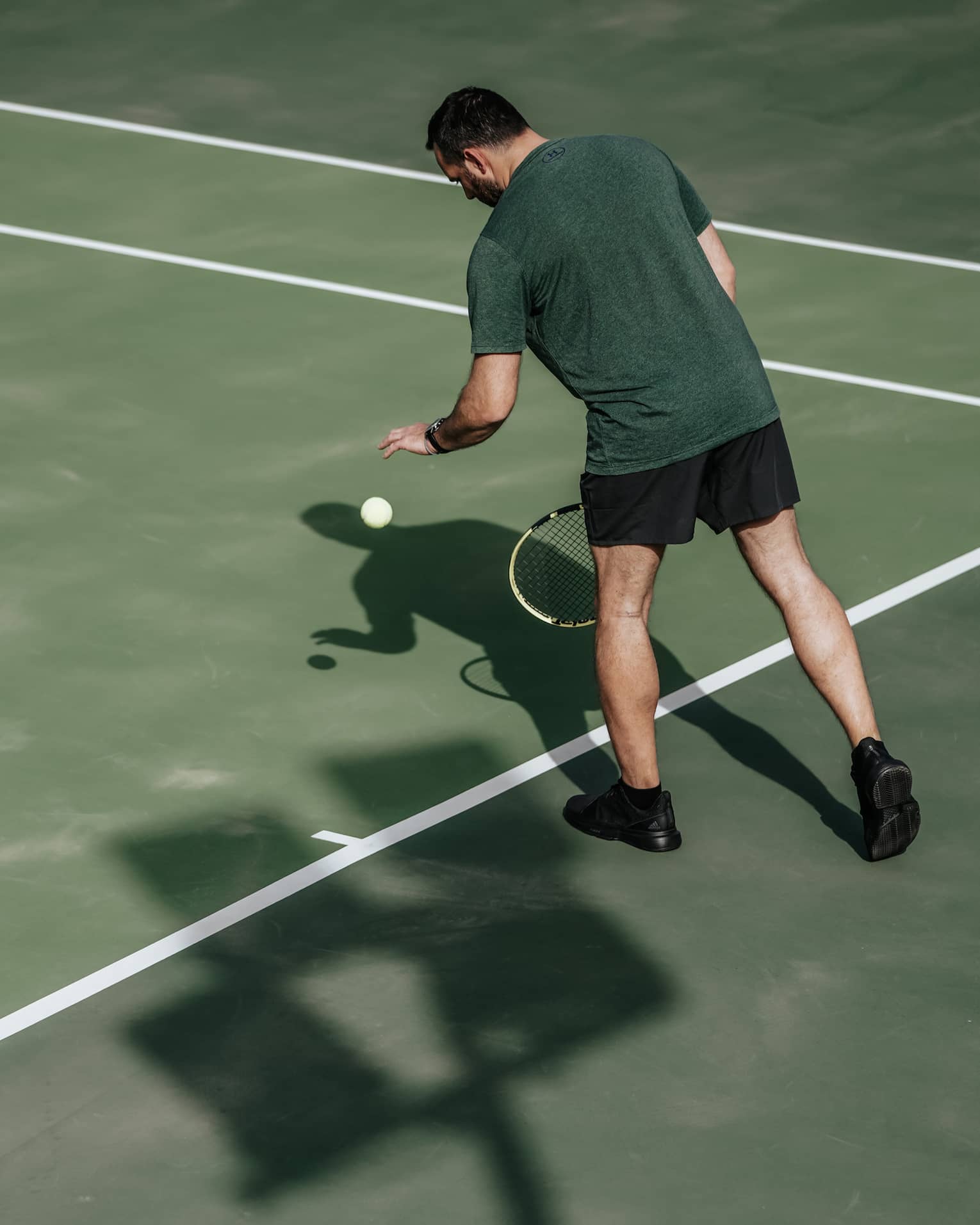 Man on tennis court bouncing tennis ball with left hand, preparing to serve