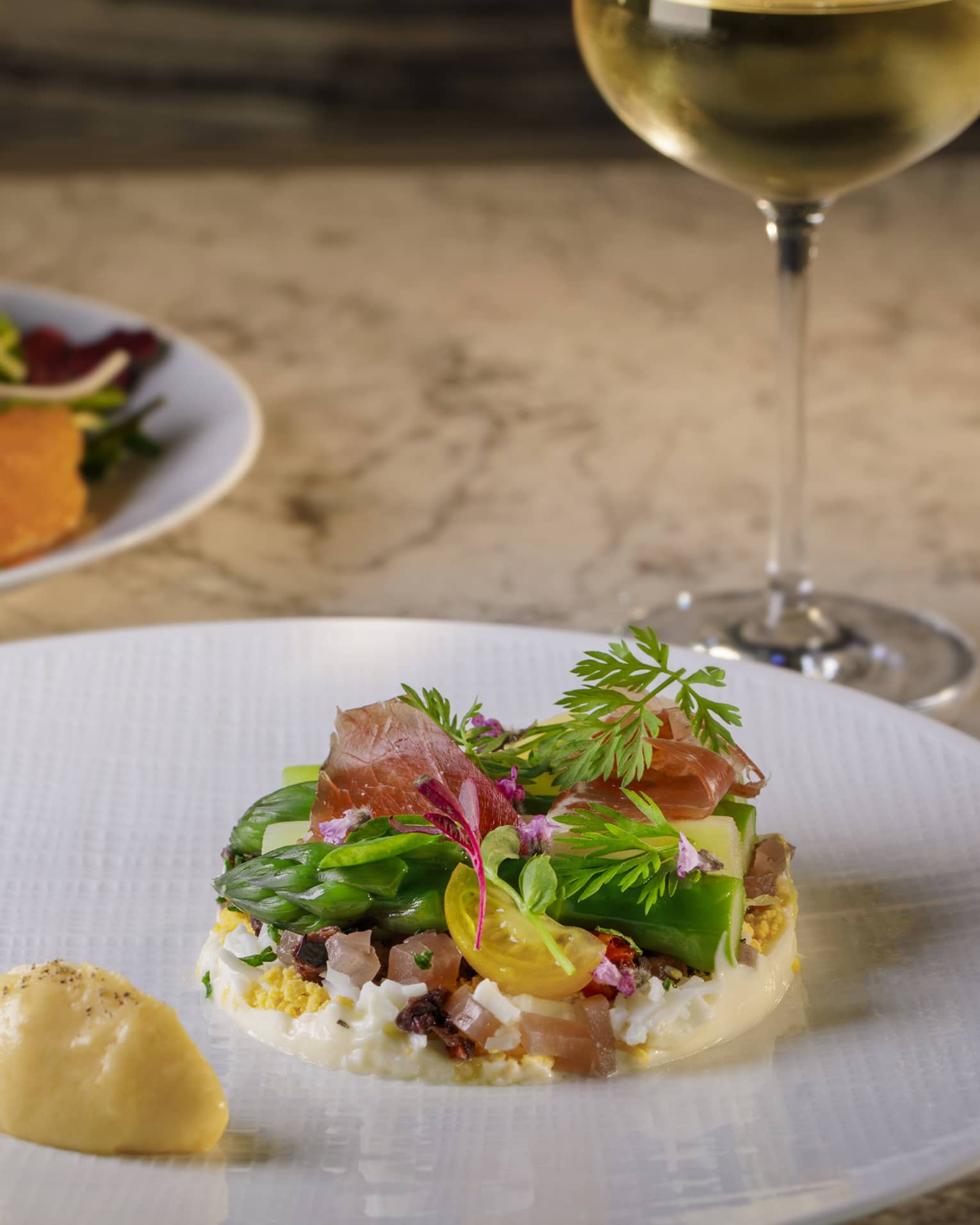 Poached egg under with green and white asparagus spears garnished with prosciutto and herbs, served with side salad and wine.