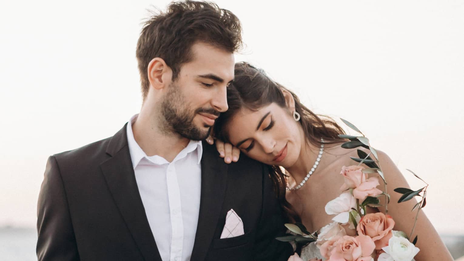 Bride holding pink rose bouquet rests head on groom's shoulder near water 