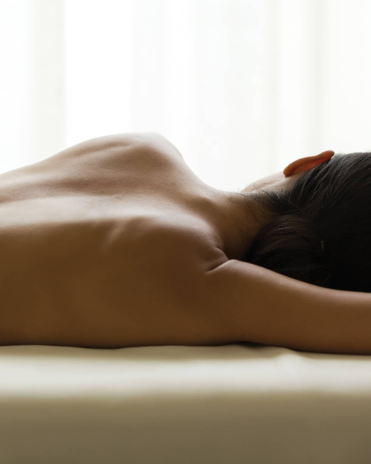 Woman's bare back as she lays on massage table under window, white curtains