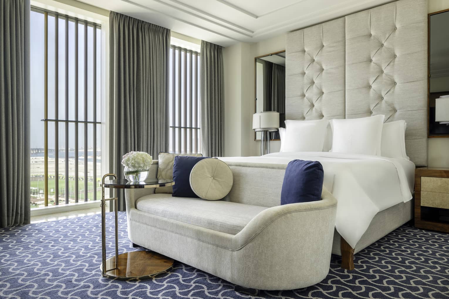 Hotel room with king bed, beige chaise at the foot of the bed, patterned carpet, ceiling-height upholstered headboard and wall of windows