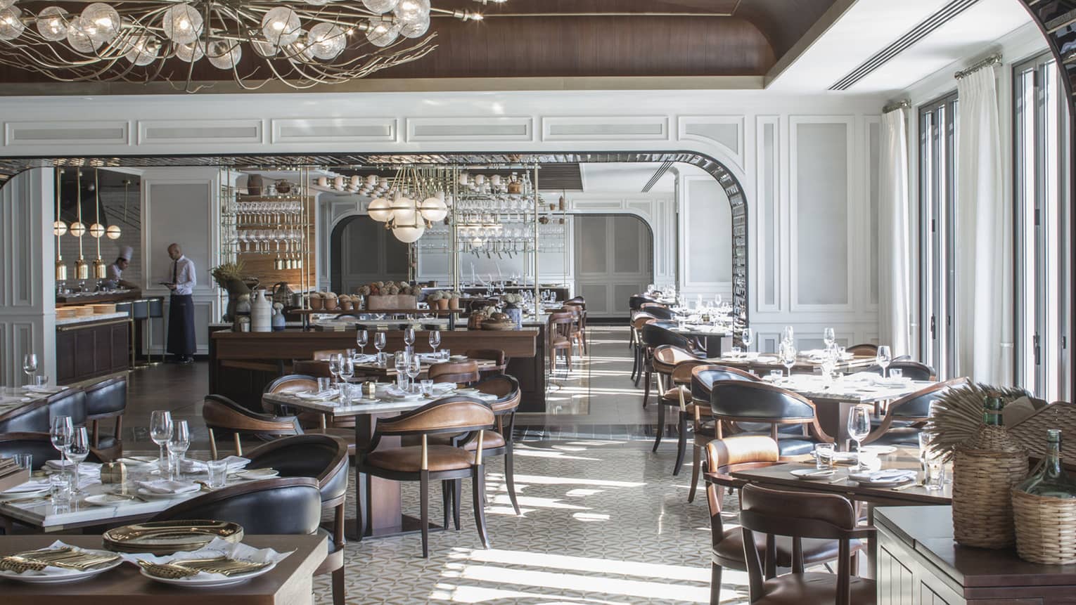 Large, sunny La Capitale dining room with elegant brown leather chairs, tables, white tile, modern chandeliers, server at counter