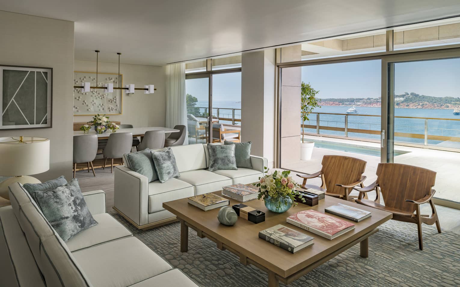 Arion Royal Suite living area with white sofas, square coffee table, arm chairs, dining table in background, floor-to-ceiling window with view of plunge pool and Mediterranean Sea