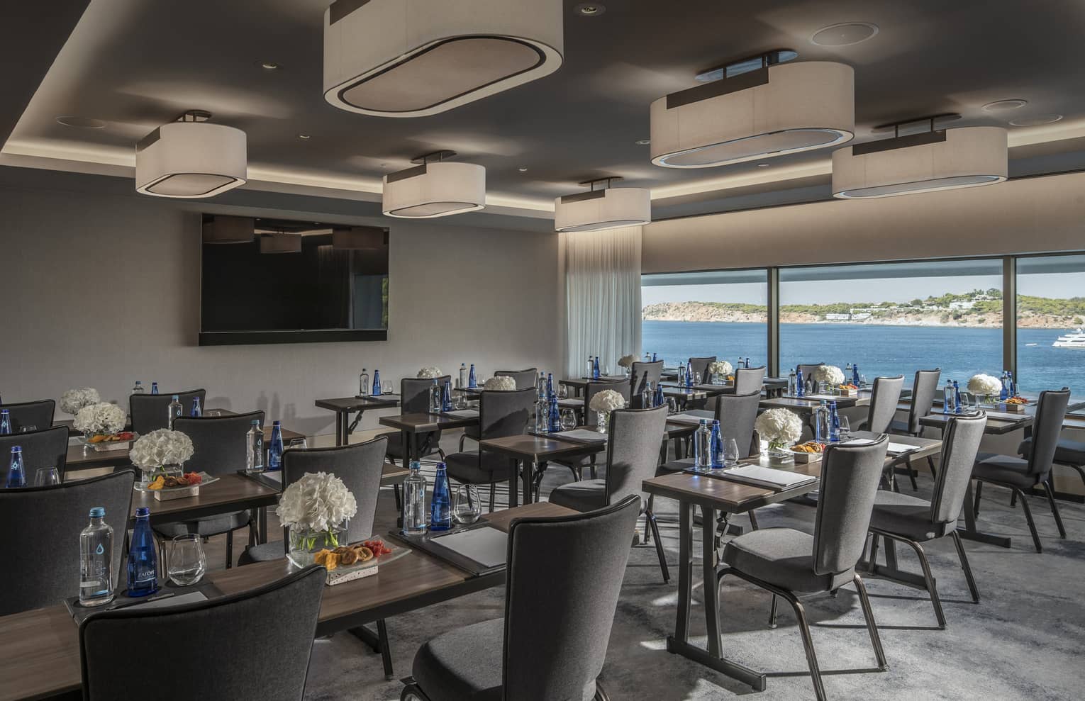 Rows of desks with water, flowers, snacks and grey chairs facing TV with ocean view