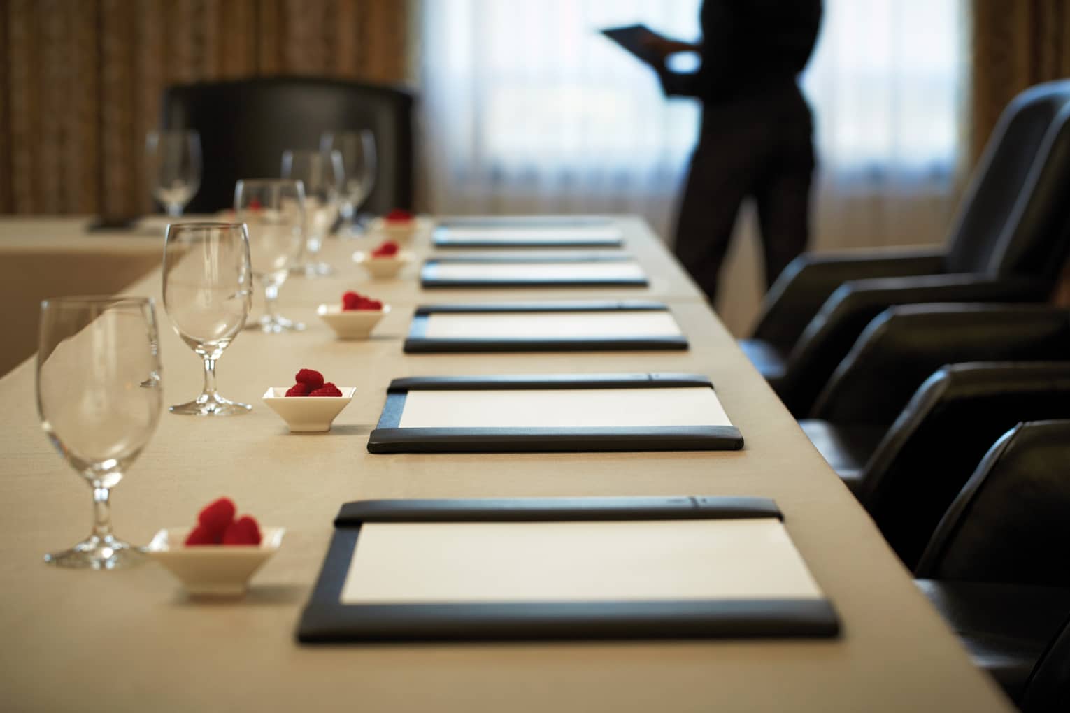 Rectangular meeting table with six place settings and a person standing in the background holding a tablet 