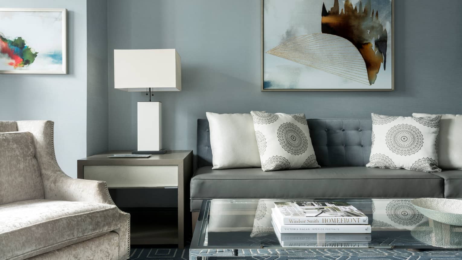 Midtown Suite long grey sofa with white accent pillows below abstract painting, table, lamp, armchair