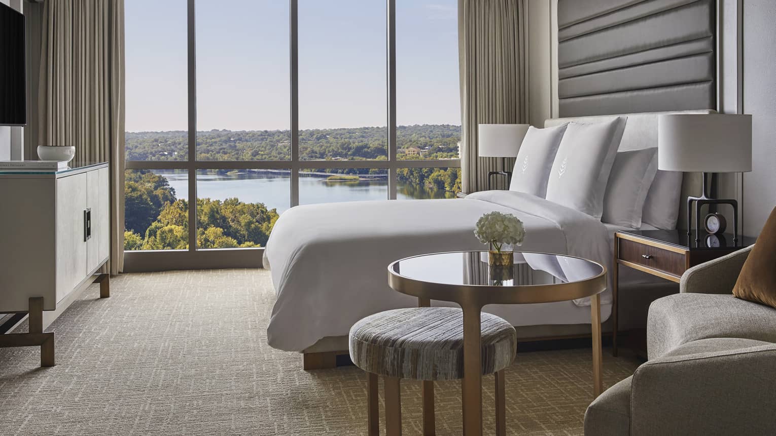 Side view of small brass and glass table, hotel bed by floor to ceiling window and lake views