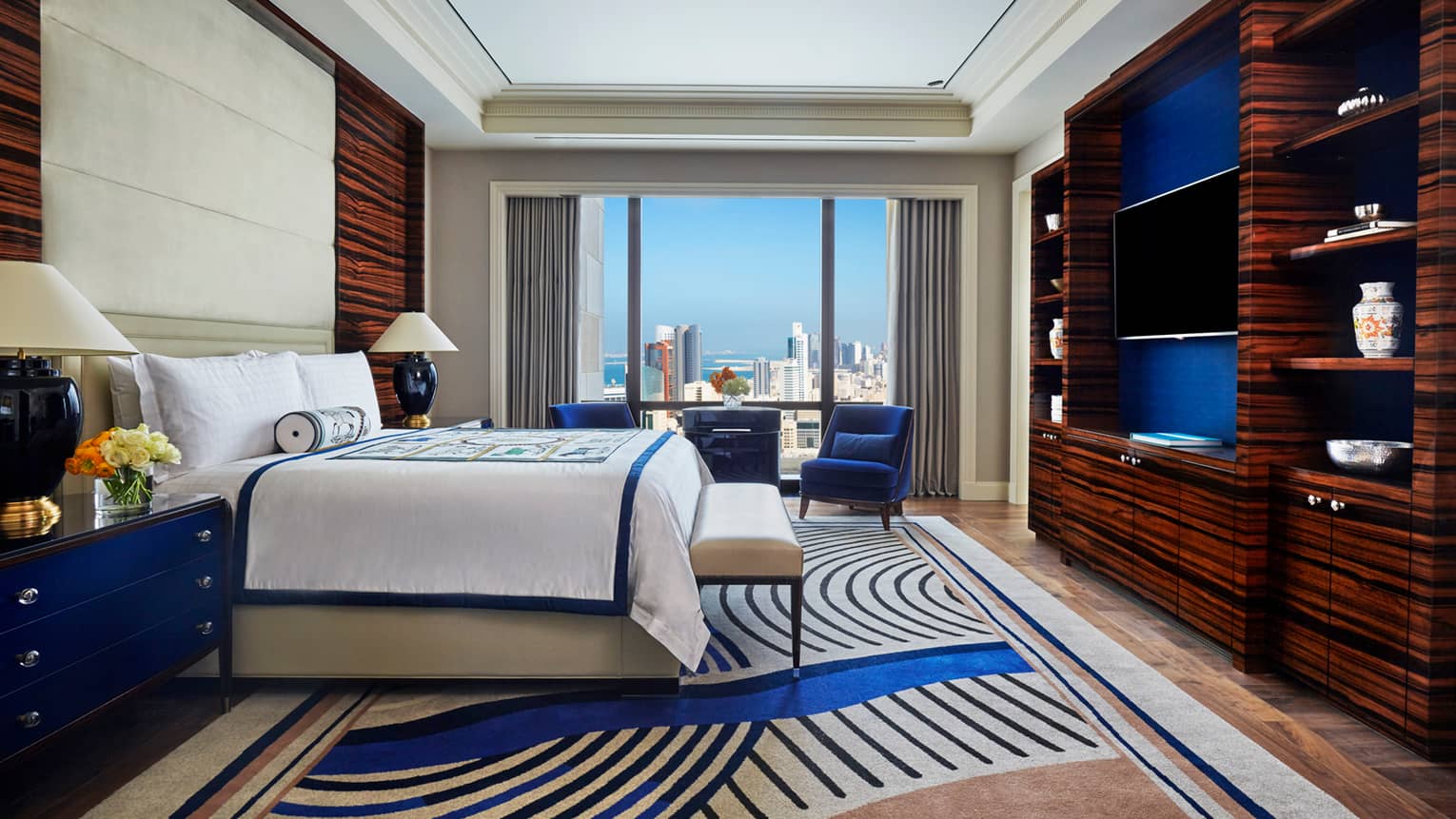 Superior Room side view of bed with decorative blanket and rolled pillow, modern blue carpet, armchair by window