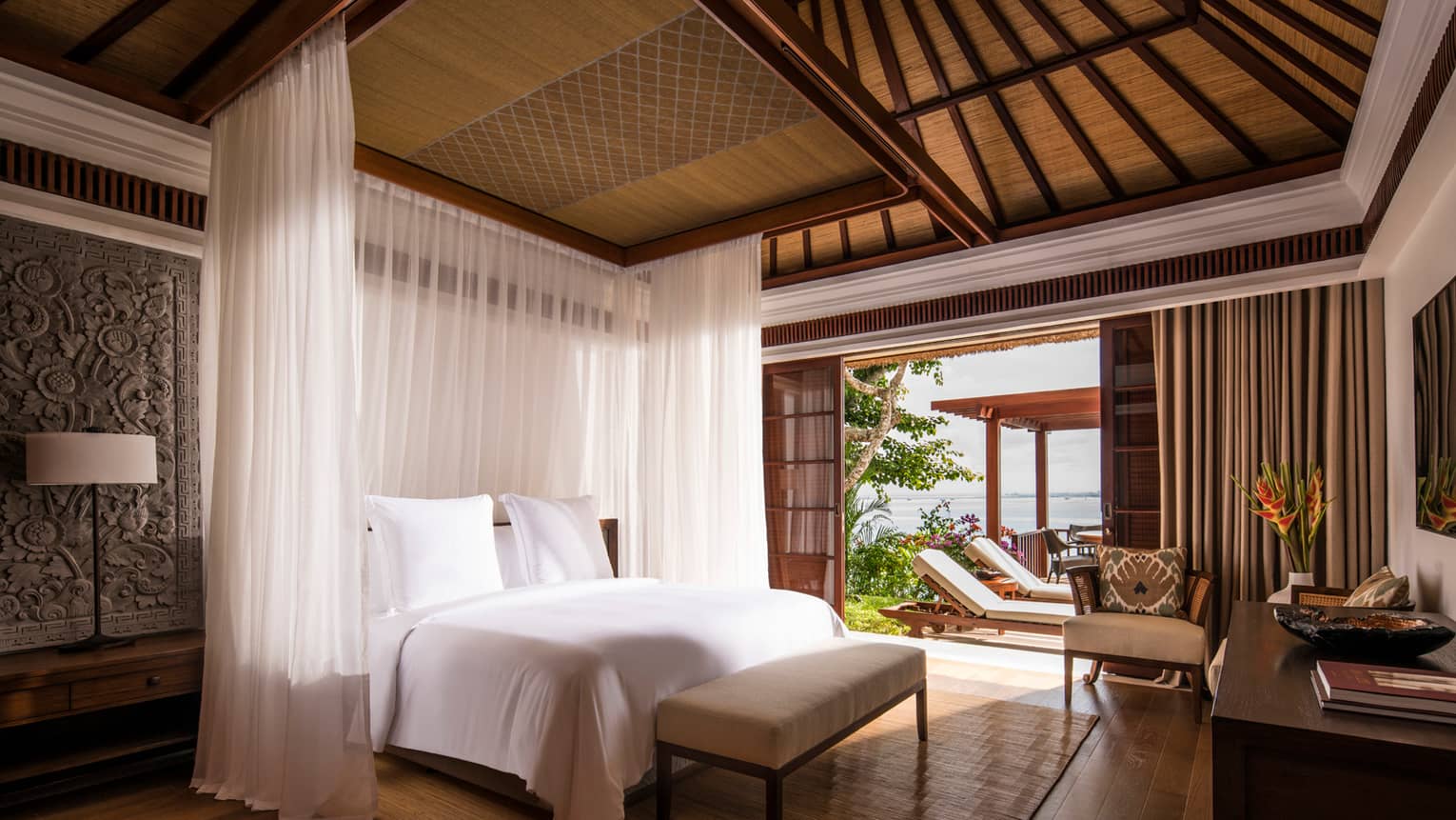 Villa bed with sheer white curtains hanging around each side, Balinese textiles on wall, door open to sunny patio