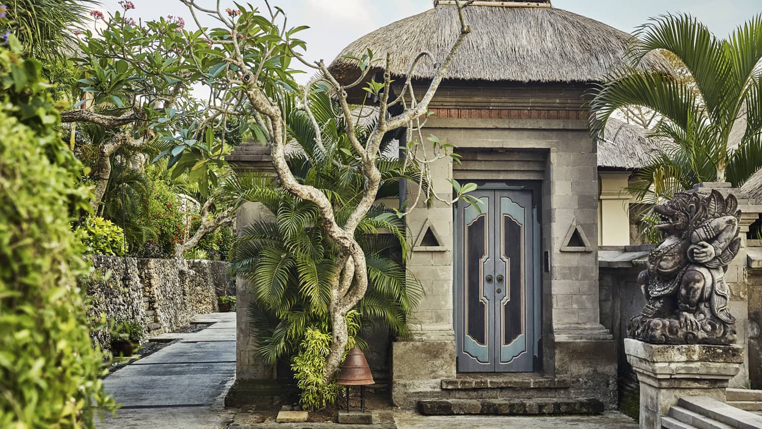 Stone wall, steps by tall rustic blue door under thatched roof, twisted tropical trees