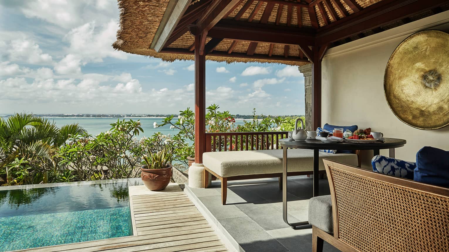 Close-up of Deluxe Villa patio corner with swimming pool, plant, white bench under thatched roof, ocean view