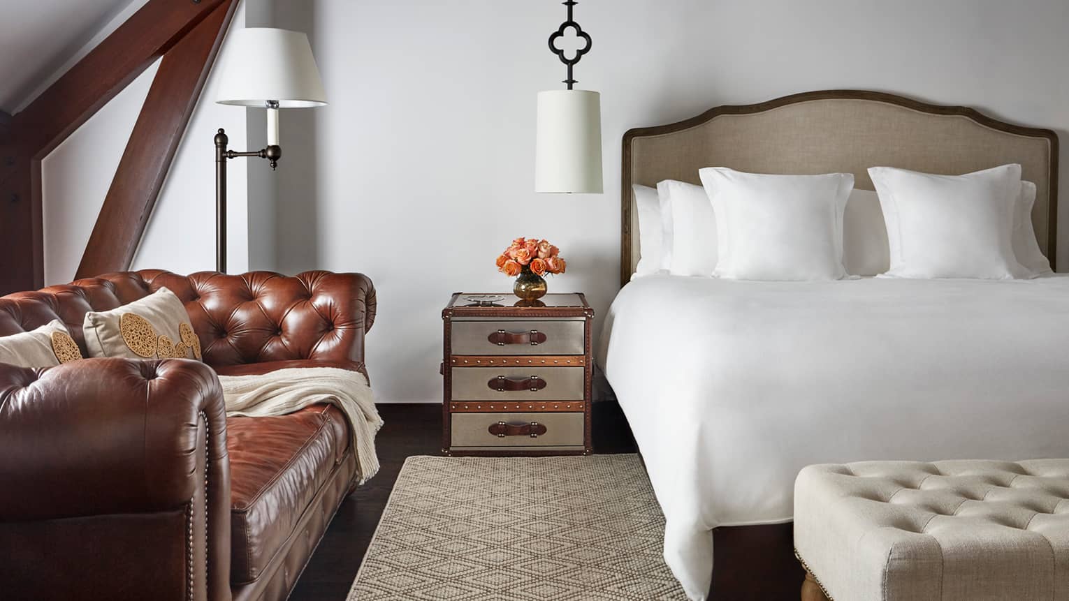 Brown period-style leather sofa under sloped beam roof next to nightstand, bed, cushioned bench