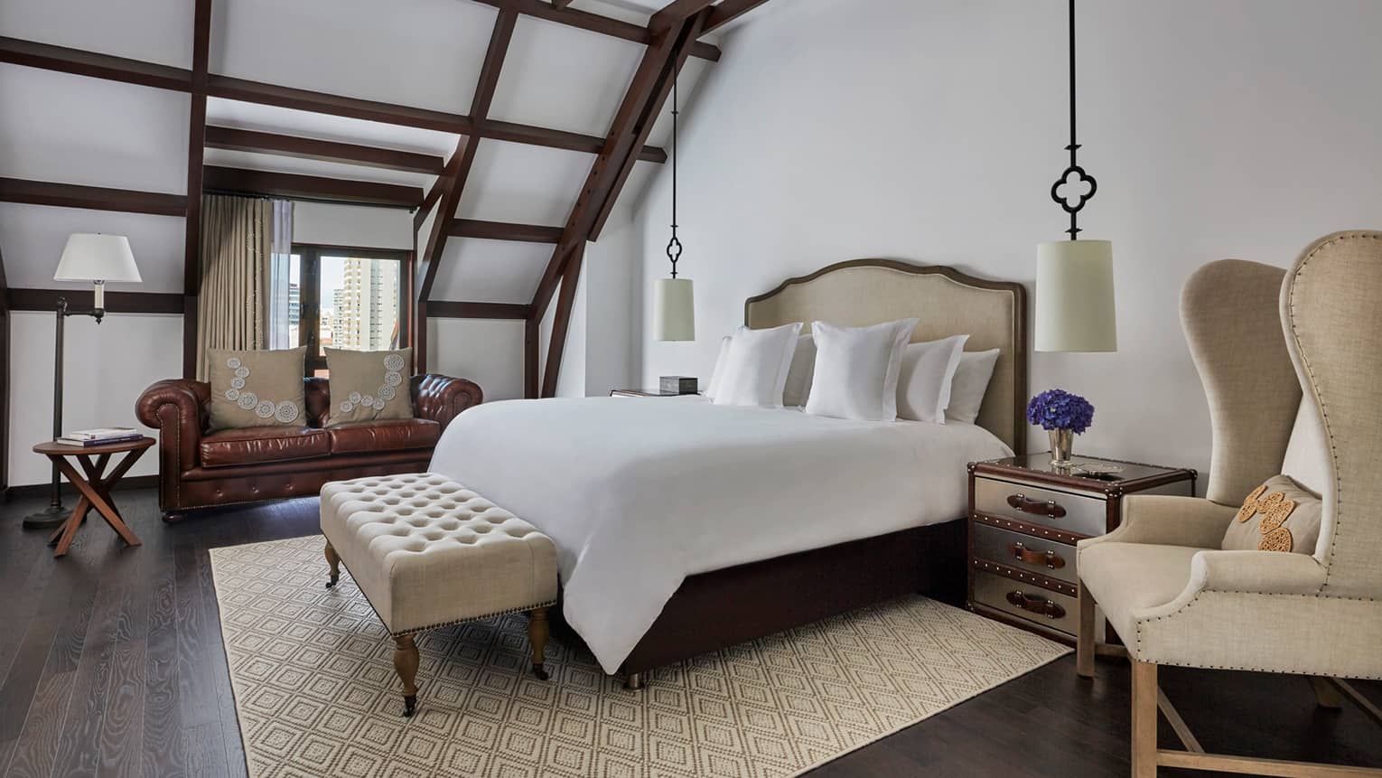 Brown leather loveseat in front of window, beamed ceilings sloped over bed, plush bench and armchair