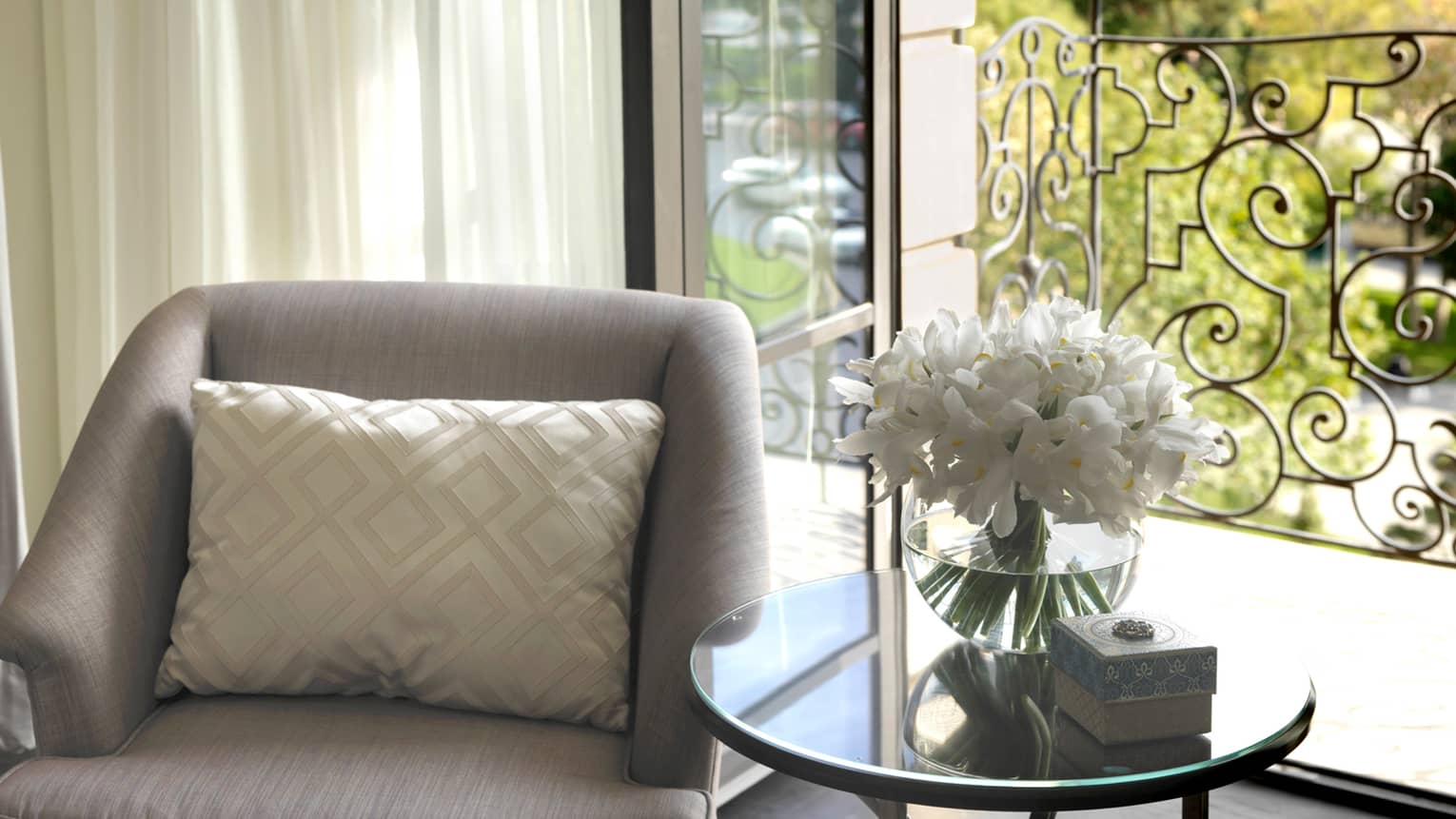Close up of grey armchair with white accent pillow, glass table and vase with white flowers in sunny window