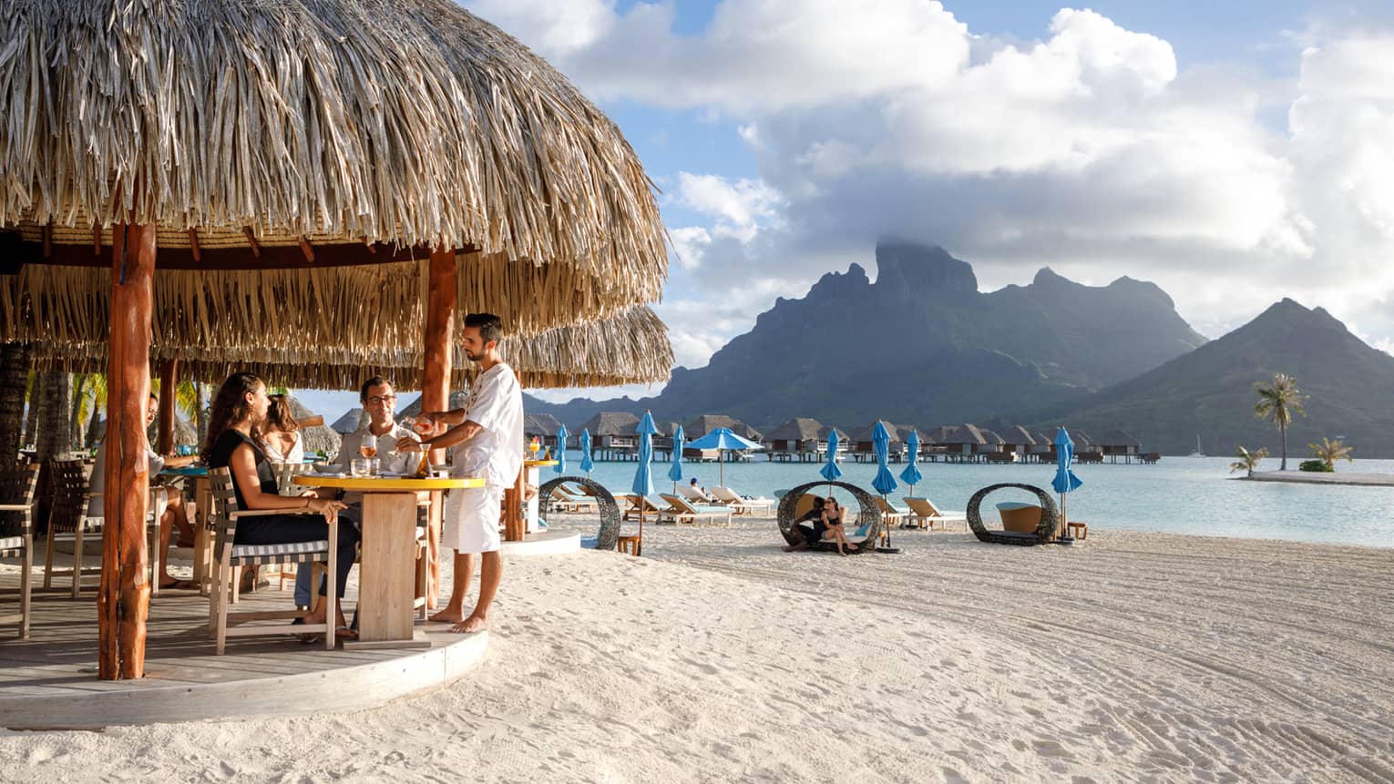 Couple seated in bar under grass roof on beach, view of people in personal cabanas, mountain view