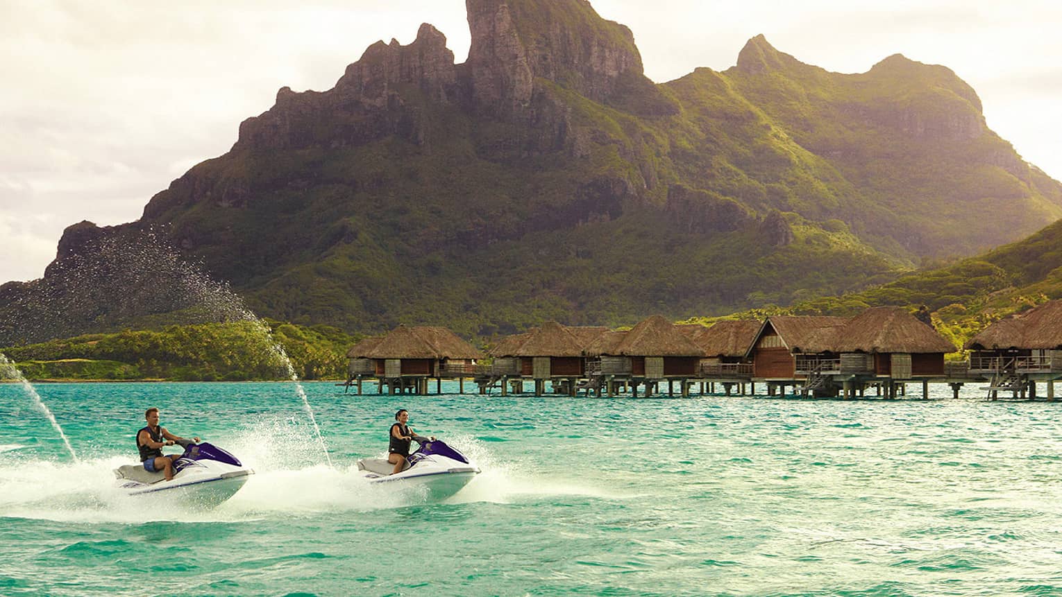 Two jet skis ride along lagoon past row of overwater bungalows, Bora Bora mountain