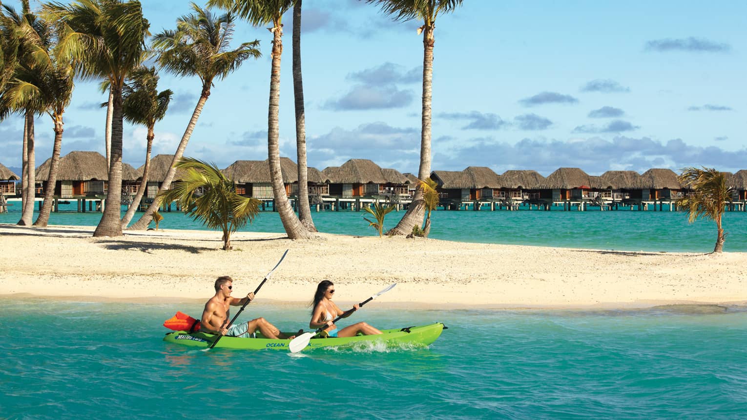 Couple kayaks past white sand motu and palm trees, underwater bungalows in background