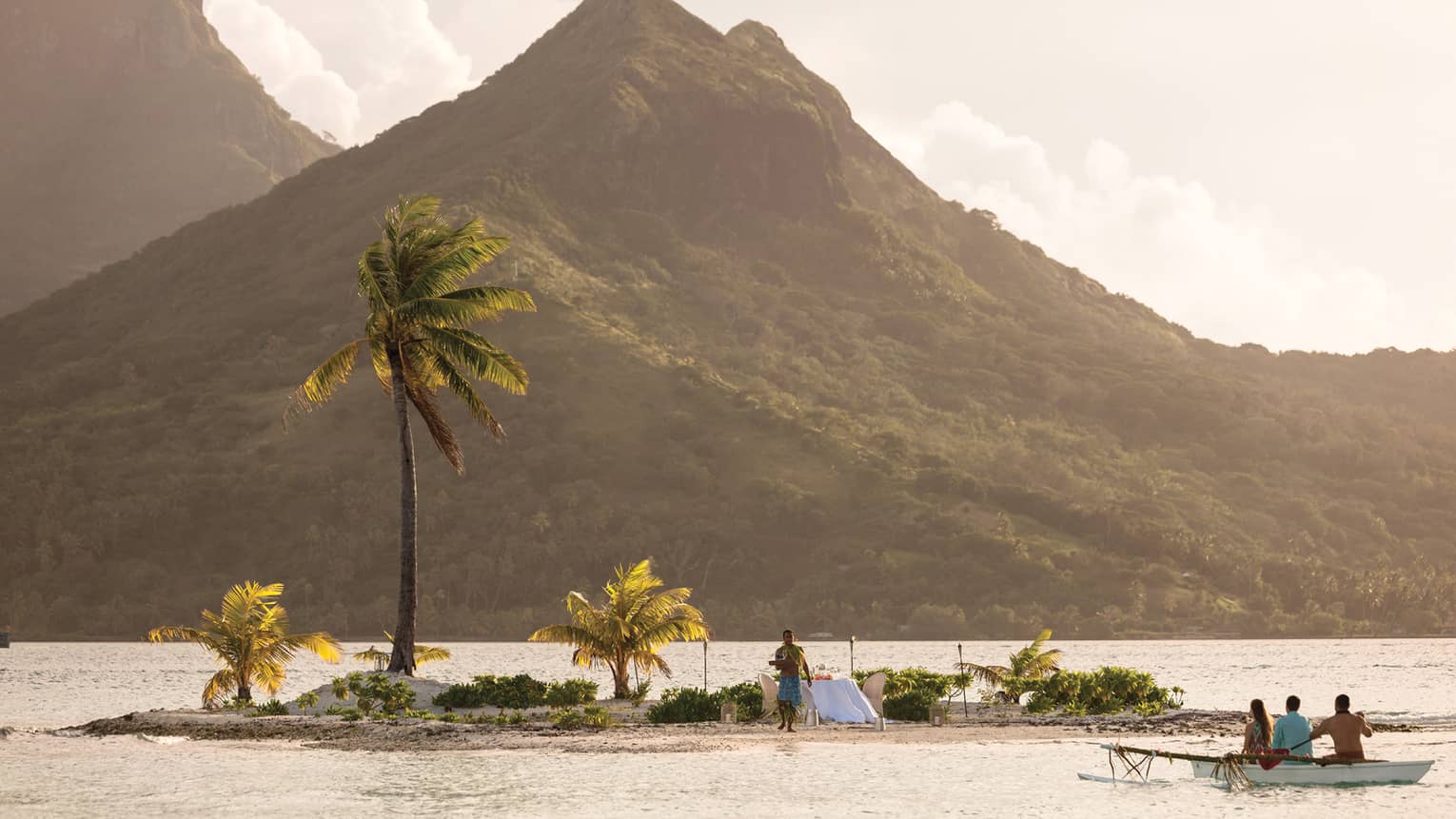 Boat approaches small private motu island at sunset where dining table with white cloth and server await