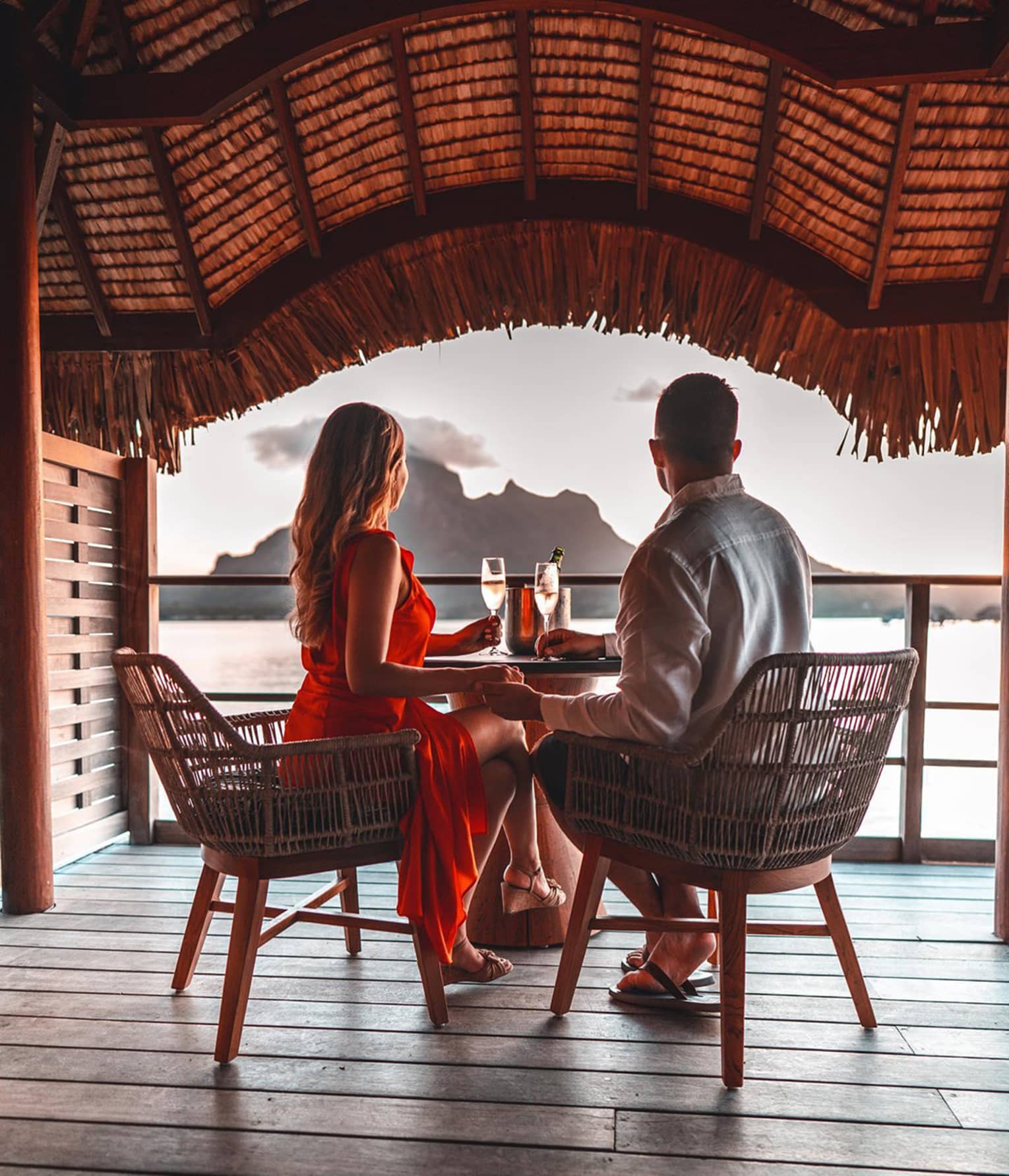 Two people enjoying a sunset view from a waterfront deck with drinks, overlooking a mountain and ocean