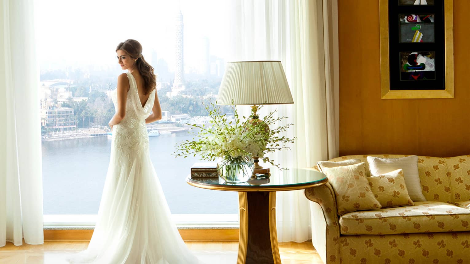 Bride wearing elegant long wedding gown stands at sunny window in hotel suite