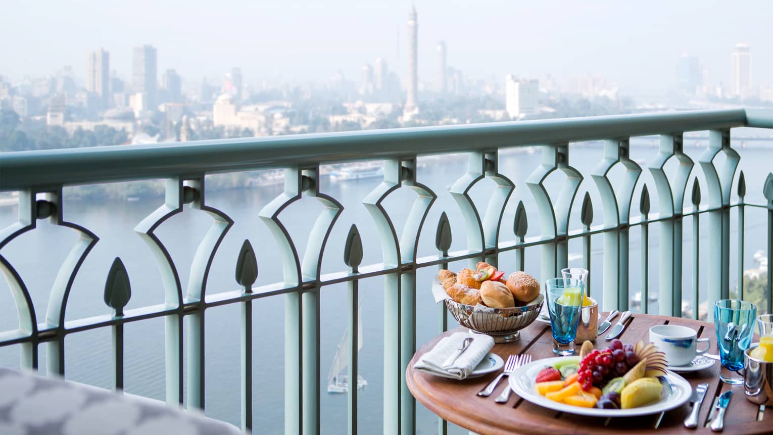 Close-up of balcony table with plate of fresh fruit, bread and orange juice, river and city view below